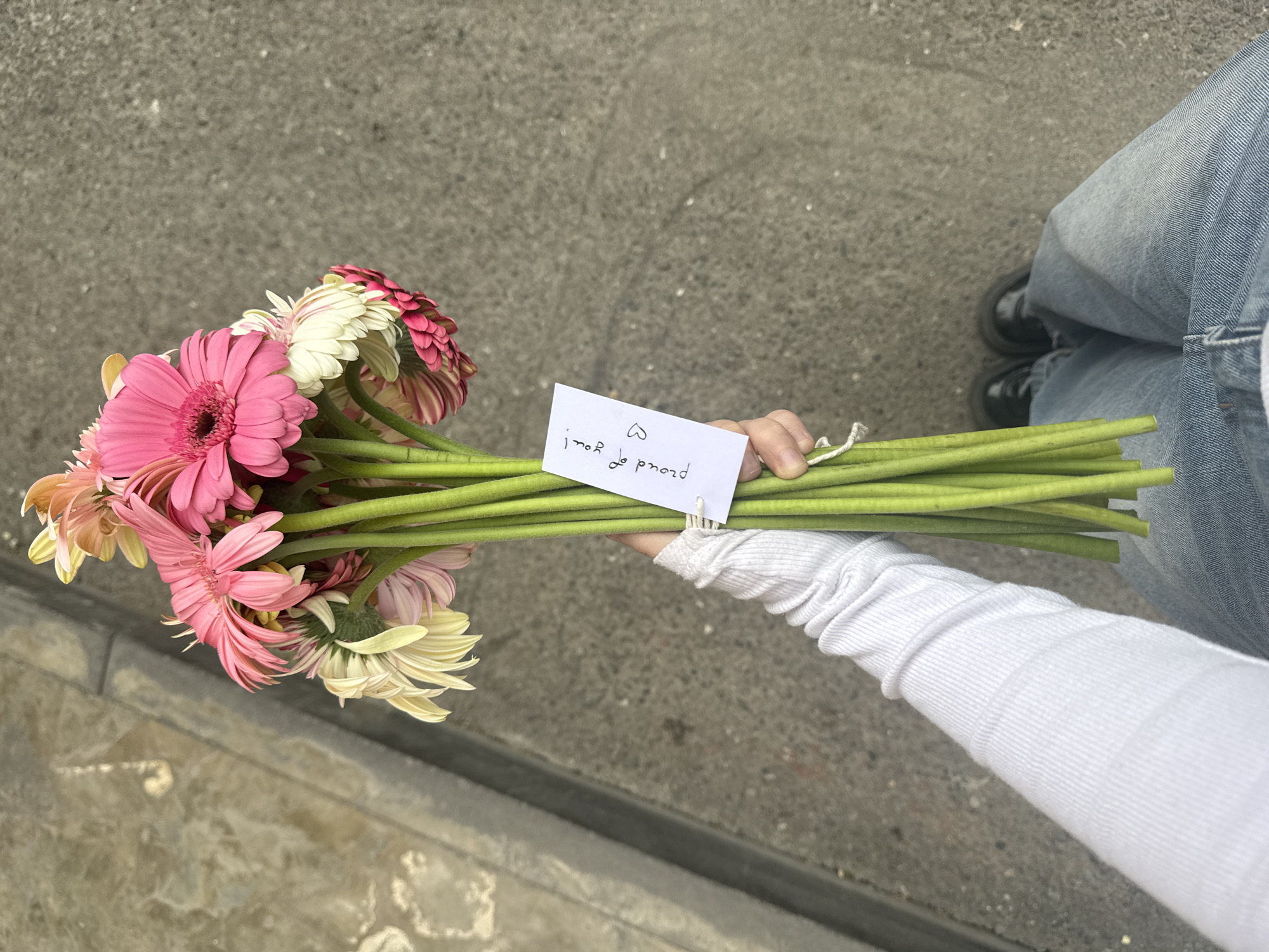 A top-down medium shot shows a person holding a bouquet of Gerbera daisies with a small white card. The flowers are arranged with pink and white blossoms, and the long green stems are gathered together. A small white card is attached near the base of the stems, bearing a handwritten message. The person holding the flowers wears a white long-sleeved shirt and blue jeans. Black shoes are visible in the upper right. The background consists of a gray concrete surface, possibly a sidewalk. The location is not discernible as Yerevan, Armenia.