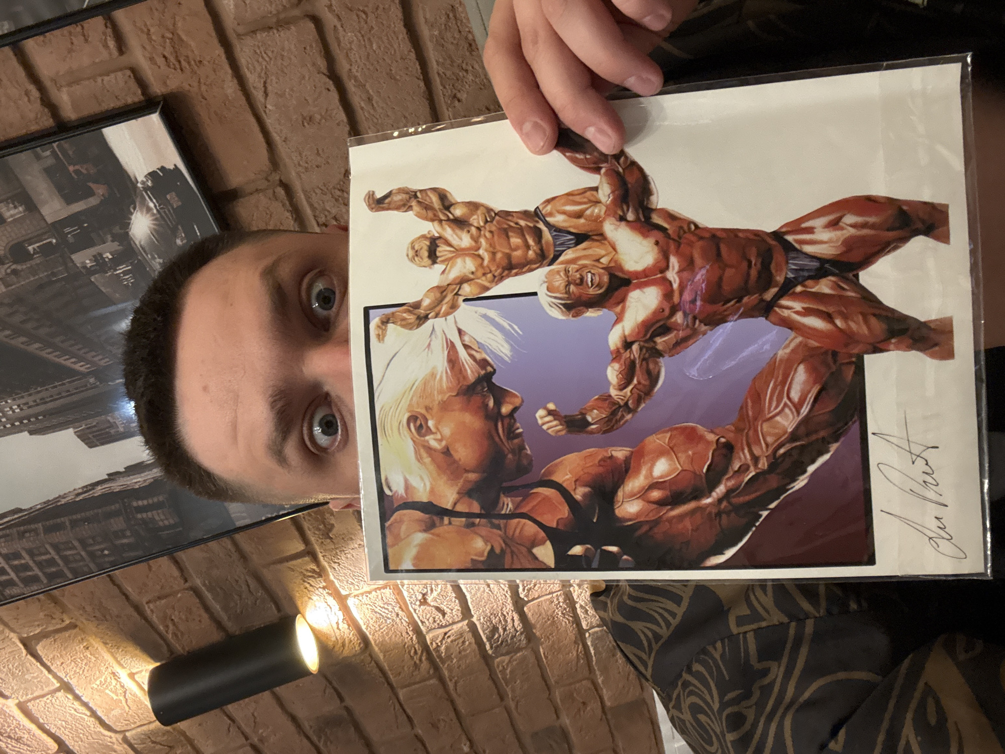 A Caucasian man with dark hair and wide eyes is holding up a comic book or print with an image of two muscular figures engaged in what appears to be a wrestling match. The man's head is visible on the left side of the frame, partially obscured by a framed photo hanging on the wall behind him. The background includes a brick wall, a cylindrical black light fixture, and a patterned garment worn by the person holding the artwork. There is no mention or evidence of the location being in Vladivostok, Russia.