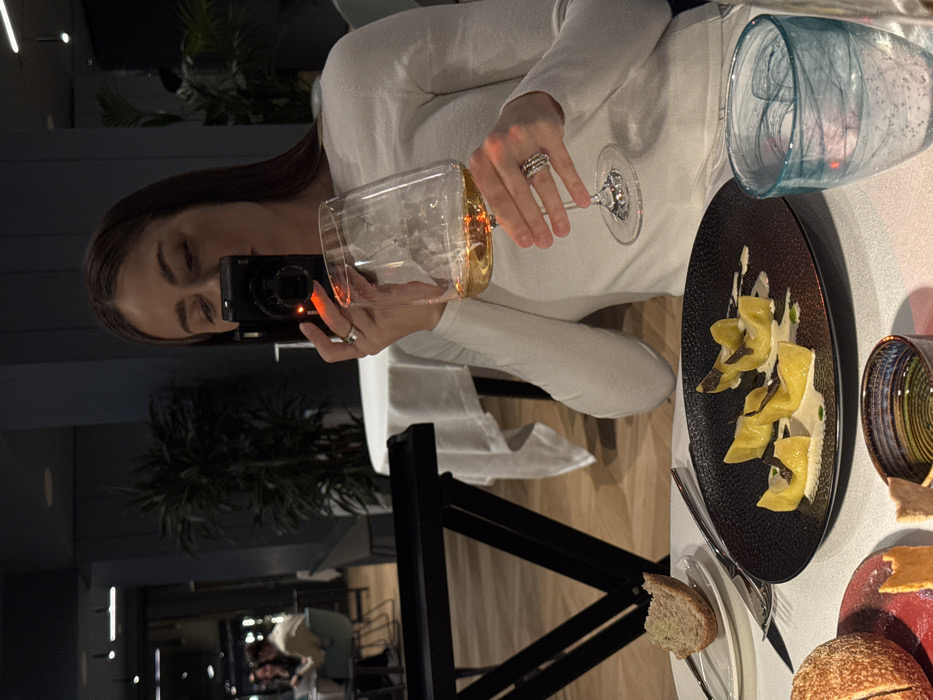 An upscale restaurant in Torbole, Italy, is depicted in a dimly lit indoor scene. In the foreground, a woman with long brown hair, wearing a white long-sleeved top, is seated at a table covered with a white tablecloth. She holds a black compact camera, exhibiting a red indicator light, to her face with her right hand, seemingly capturing a photograph. Her left hand holds a clear wine glass filled with light amber liquid. Multiple rings are visible on her fingers.
On the table in front of her, a black textured plate presents five pieces of yellow folded pasta, arranged on a light sauce and garnished with dark slices, likely truffle. To the left of the pasta plate, a small white plate holds a piece of white bread with a bite removed, alongside a knife and fork. A dark bread roll rests on a red-patterned plate. A small, dark-rimmed bowl contains a clear liquid, possibly olive oil, and a small food item. To the right, a clear glass with a distinctive light blue swirl pattern contains ice and water.
The background reveals a modern dining environment with dark blue walls, light wooden flooring, and several potted plants. Another white-clothed table is visible with two indistinct figures seated, appearing to be dining. A black serving trolley or stand is partially visible on the left side of the frame.