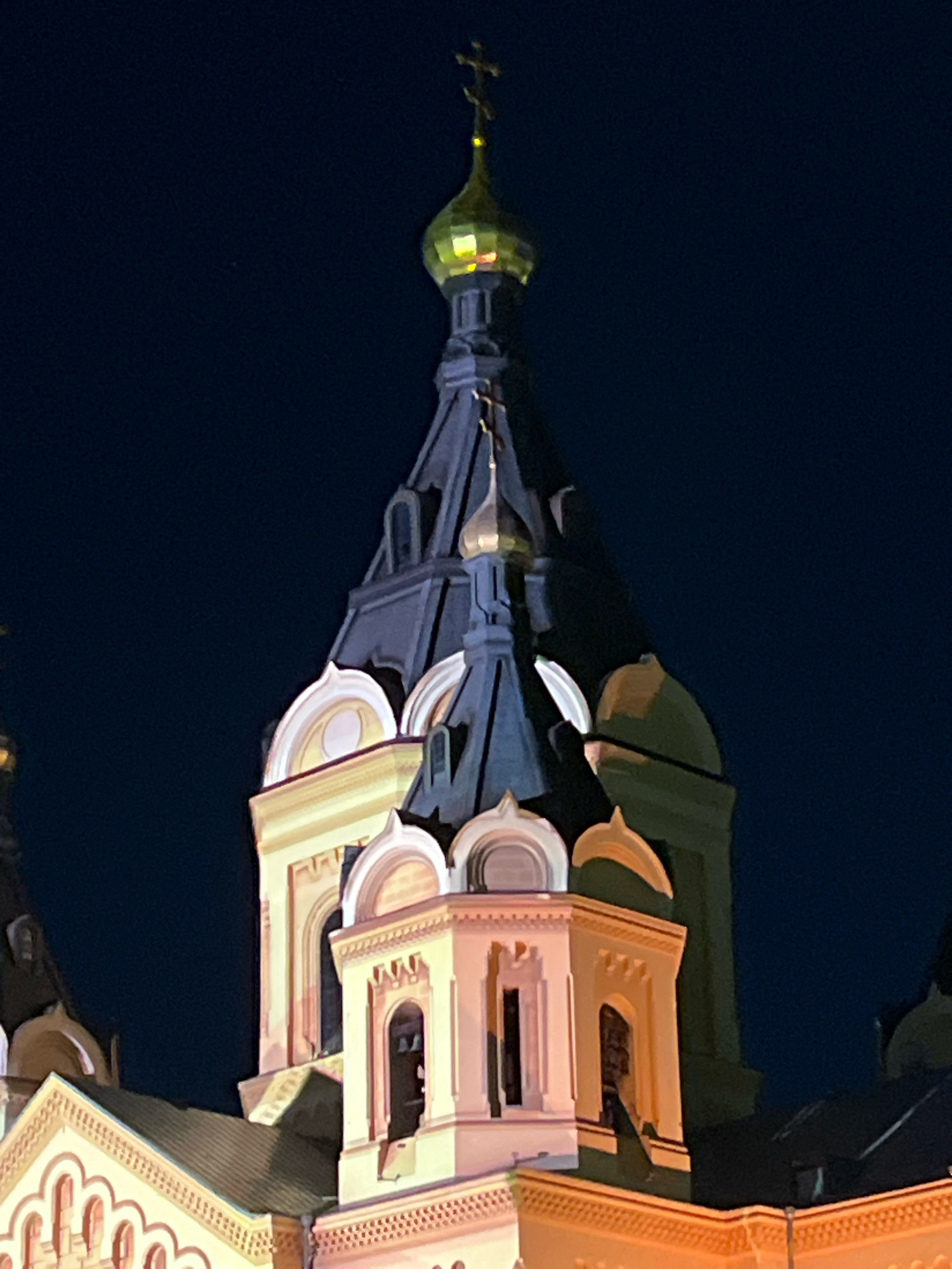 The image shows the illuminated bell tower of a church in Nizhny Novgorod, Russia, at night. The dominant structure is a tall, multi-tiered bell tower with a dark, sloped roof, topped with a golden, onion-shaped dome bearing a cross. The tower is predominantly yellow with white trim and features arched windows. Adjacent to the bell tower, other architectural elements of the church are visible, including arched rooflines and smaller domes. The sky behind the church is dark, indicating nighttime. The lighting suggests artificial illumination of the church structure.