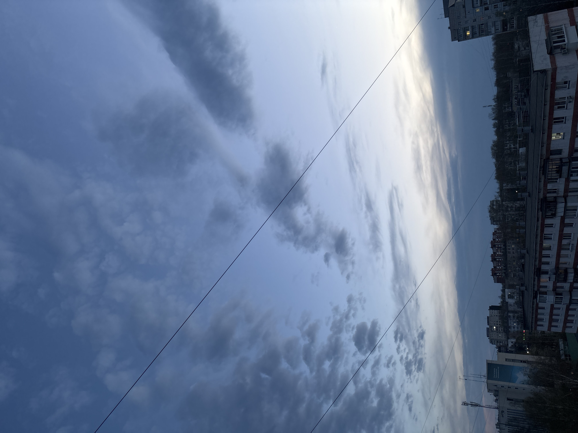 The image shows a dusk or dawn scene in what appears to be an urban area, possibly Chaikovsky, Russia. The sky is filled with clouds, ranging from dark and dense to thin and illuminated by the sun's rays. Overhead power lines are visible diagonally crossing the frame. Buildings, likely residential apartments, are located on the right side of the frame. Some windows are lit. Trees are visible between the buildings. A sign is attached to one of the buildings. The camera angle is looking upwards, with the horizon on the right side of the frame.