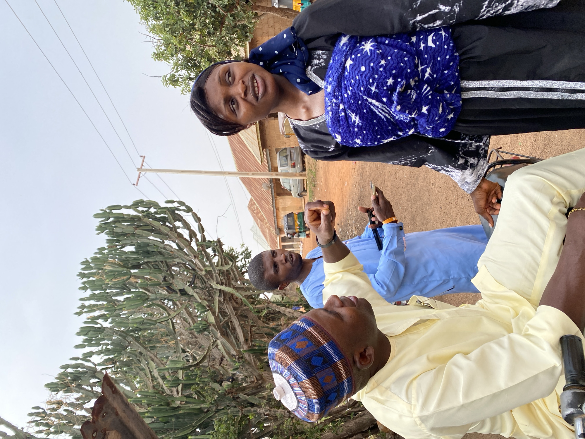 The image appears to be taken outdoors in Kuru Karama, Nigeria. The scene includes three individuals. A woman in a black and white dress with a blue scarf is visible in the upper portion of the image. Further down, two men are seen. One man wears a light yellow shirt with a blue and brown cap and is partially seated on a wheeled object, possibly a wheelchair or vehicle. Another man in a light blue shirt is standing next to him, holding a mobile phone. Behind the men, there is a large, leafy tree. In the background, a building and a white vehicle are visible. Power lines are above the buildings. The ground is dirt-colored. The image is taken at a slightly tilted angle.