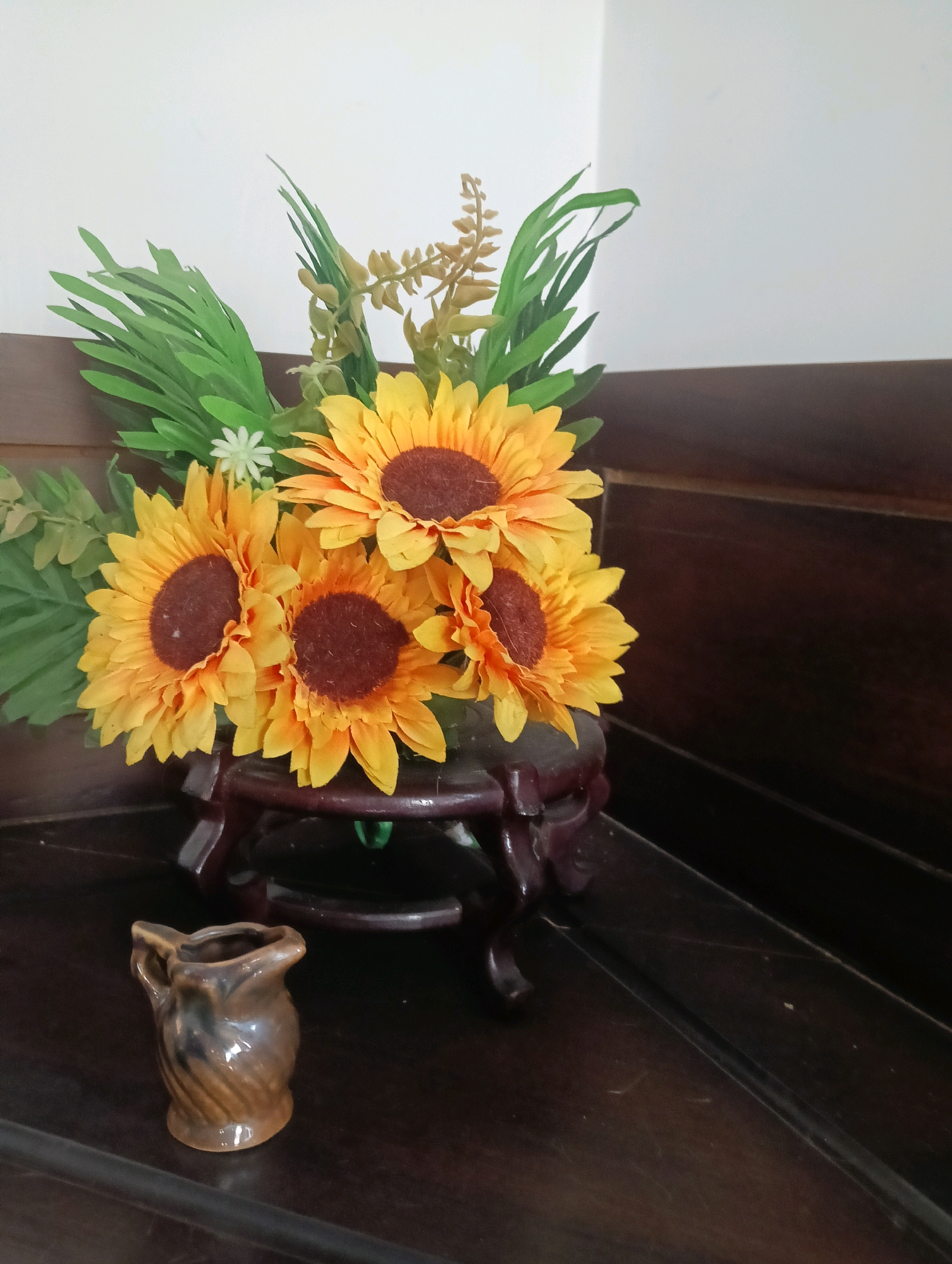 A still life arrangement features artificial sunflowers with dark centers and yellow petals, accented by green artificial foliage. The bouquet is displayed on a dark, ornate wooden stand. To the left of the stand, a small, brown ceramic jug with a glossy finish and a twisted texture sits on a dark wooden surface. The background consists of a white wall and dark wooden paneling. This scene is likely located in a domestic setting within Kromengan, Indonesia, possibly a living room or entryway, given the furniture and decor.