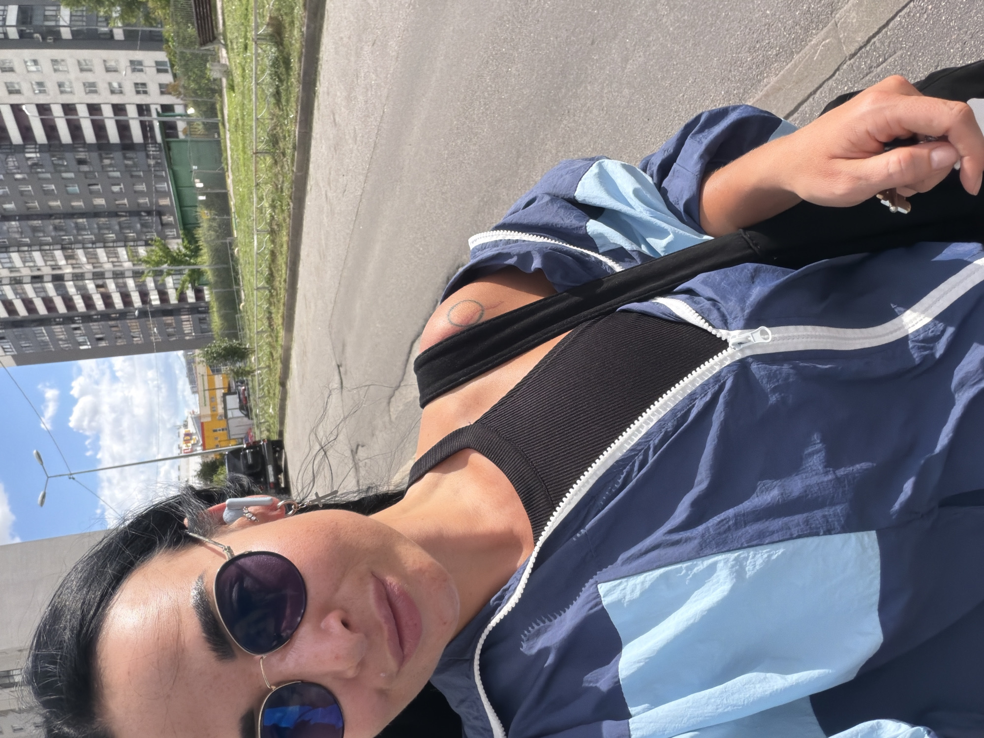 A young woman is taking a selfie outdoors in Saint Petersburg, Russia. She is wearing a blue and light blue windbreaker, a black tank top, sunglasses, and has earbuds in her ears. A black bag is slung across her body. Her left arm is visible, showing a small circle tattoo on her upper arm. The background shows a tall, modern apartment building, a street, and a clear blue sky with scattered clouds. The time appears to be daytime, judging by the sunlight.