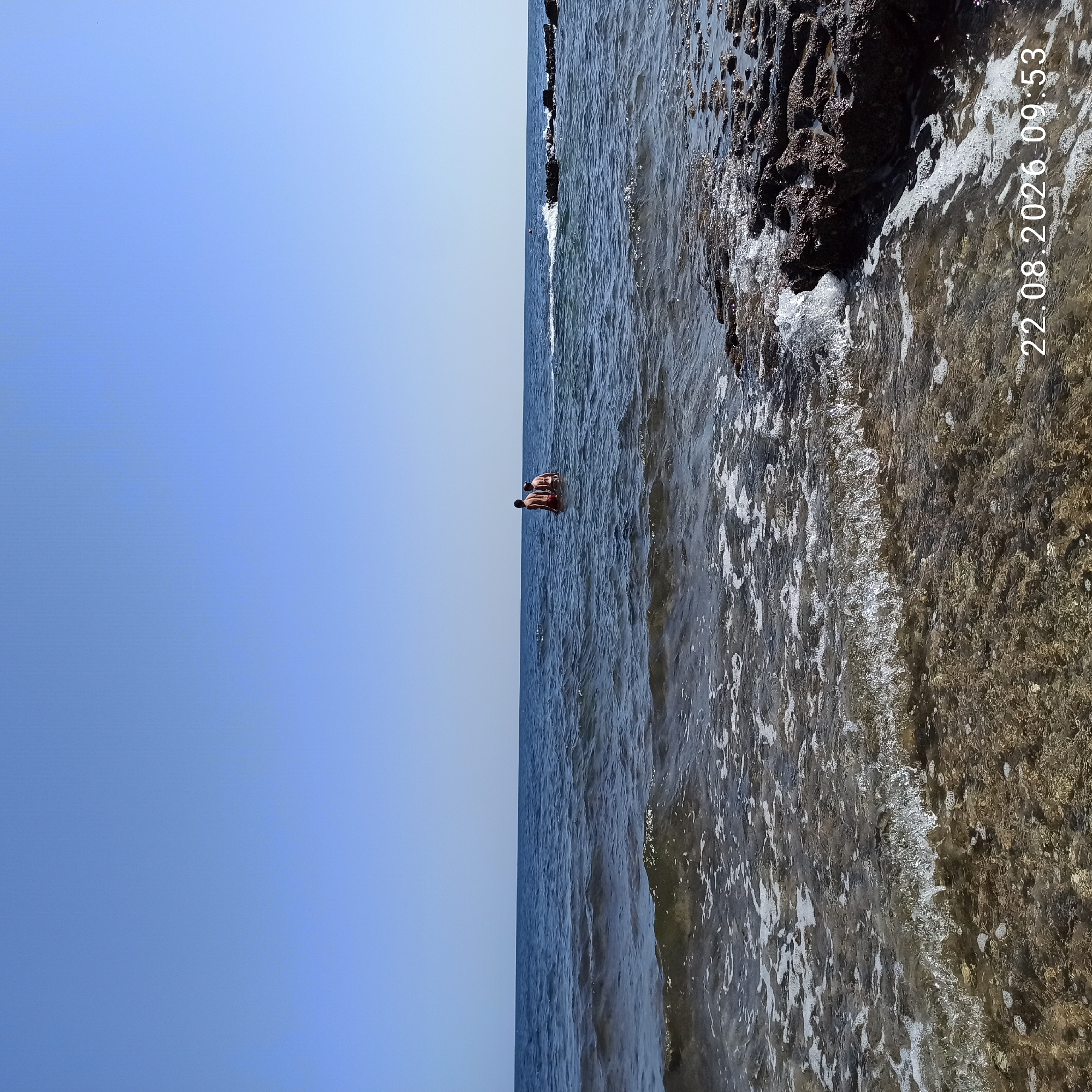 The image depicts a clear blue sky above a calm, blue ocean. In the distance, two individuals are standing in the water. The foreground shows the shallow seabed with some rocks and breaking waves. A timestamp in the bottom right corner reads "22.08.2026 09:53". The location is Molfetta, Italy.