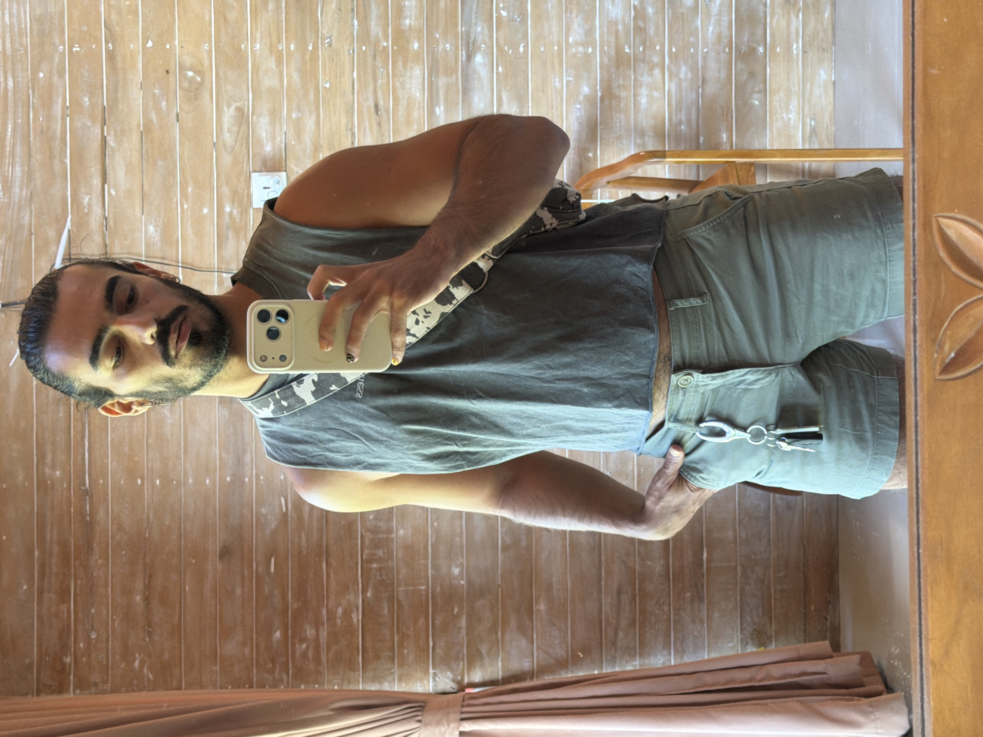 A man is taking a mirror selfie in a room with wood-paneled walls. He has dark hair tied back in a bun, a beard, and is wearing a grey tank top, green shorts, and a crossbody bag with a camouflage pattern. His left hand is in his pocket, and he is holding a smartphone in his right hand. There is a light-colored curtain on the left side of the frame and a wooden chair on the right. The scene appears to be in Sawan, Indonesia.