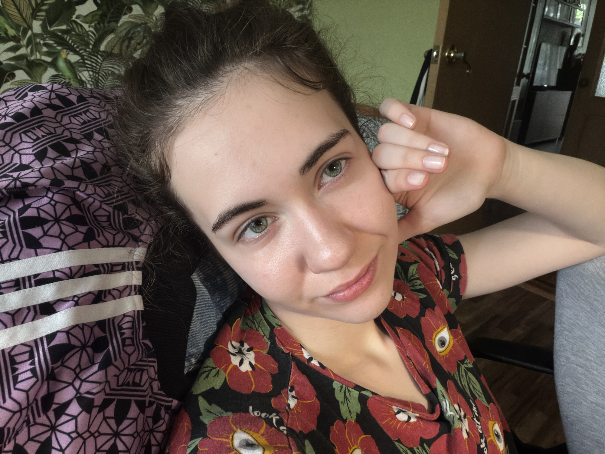 A young woman with green eyes and dark hair pulled back from her face is lying down, resting her head on her hand. She is wearing a dark t-shirt with a repeating pattern of red and yellow flowers. Her nails are clean and appear to have a light pink polish. To her left, a part of a chair with a pink and black patterned fabric is visible, featuring geometric shapes. In the background, a wall with a green, leafy wallpaper is visible, along with a portion of a wooden door with a brass doorknob. The overall impression is an indoor, casual setting. There is no information available to determine the specific location in Zavodoukovsk, Russia.