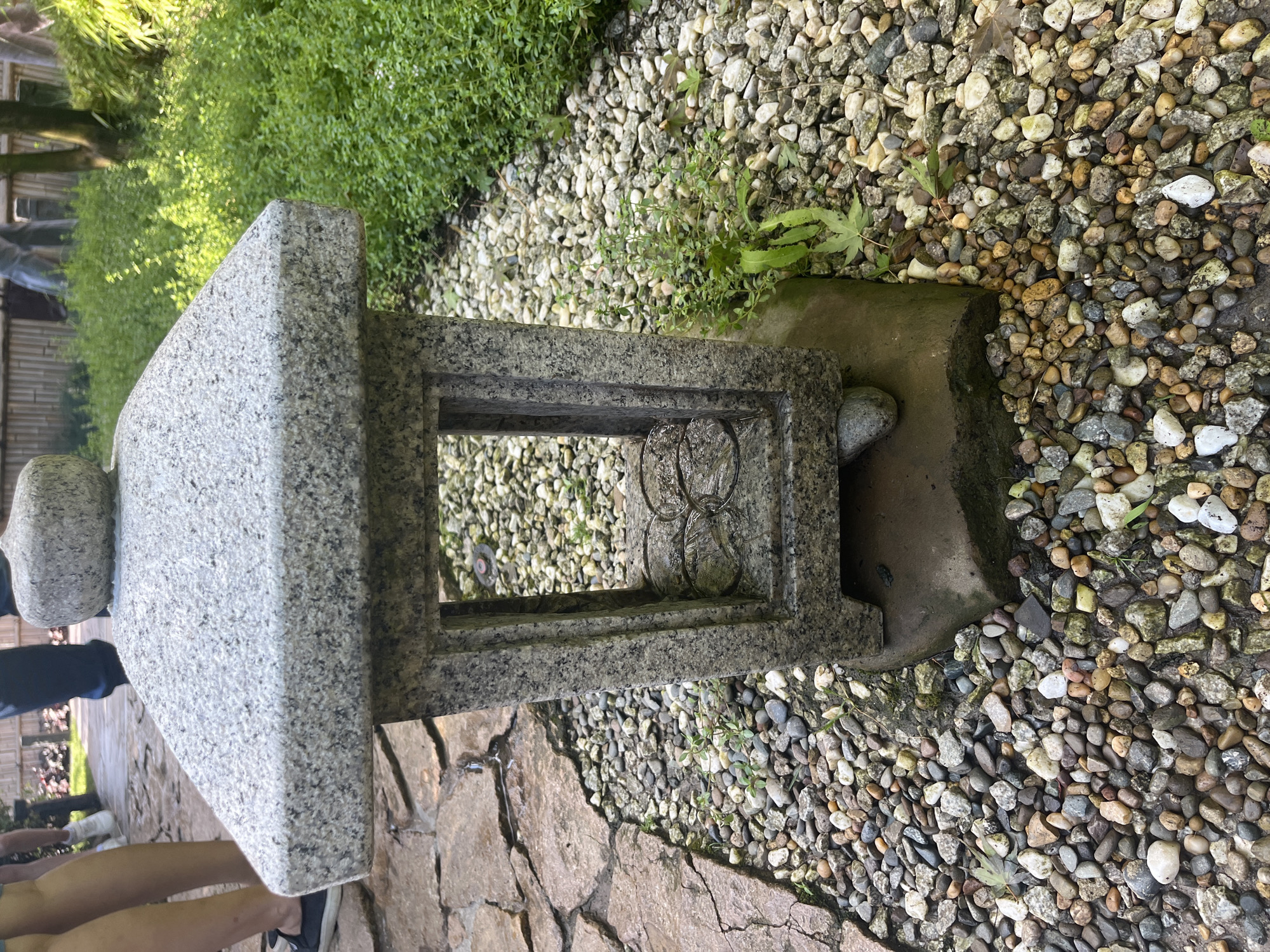 The image shows a stone lantern in a garden. The lantern is made of grey speckled granite and has a peaked roof. It is positioned on a bed of small, multi-colored pebbles, with larger flat stones forming a pathway to the left. Behind the lantern, lush green vegetation grows densely, and a bamboo fence is visible in the background. Two people's legs are visible on the left side of the frame, one wearing dark shoes and the other white sneakers, indicating they are standing on or near the stone pathway. The lighting suggests it is daytime with some sunlight filtering through. The scene is naturalistic and appears to be a well-maintained garden. There is no indication of the city of Krasnodar, Russia.