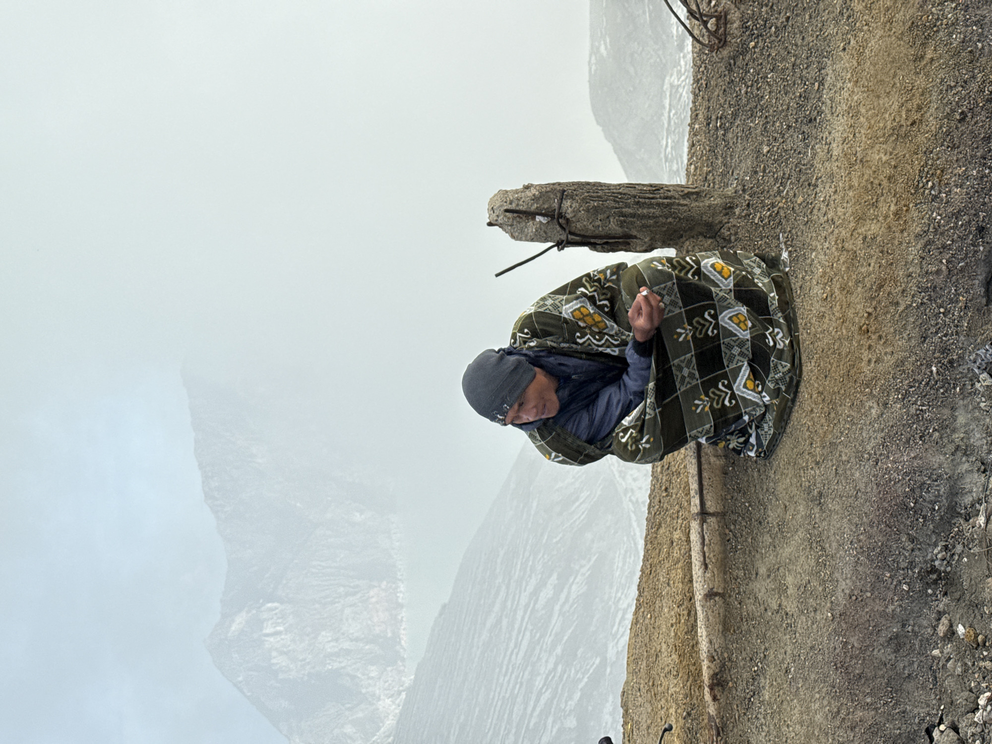 A man is wrapped in a green patterned blanket while sitting on a dirt ledge near a weathered concrete post with exposed rebar. He wears a dark jacket and a gray beanie. The background shows a foggy mountain landscape. This is in Unknown, Indonesia.