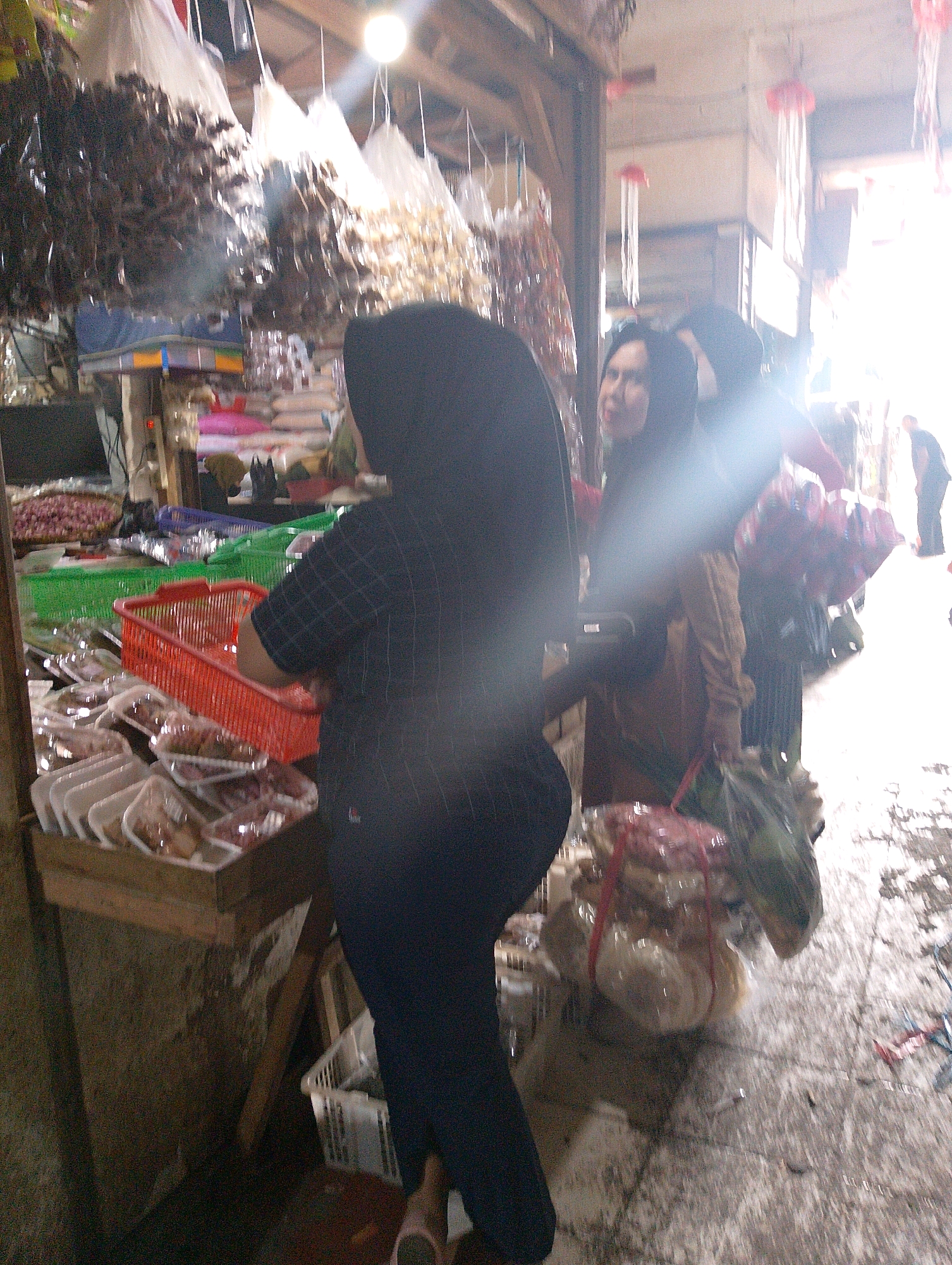 In Lembang, Indonesia, a busy market scene unfolds. Several people, predominantly women wearing hijabs, are visible. One woman in the foreground is wearing a black hijab and a black checkered shirt. Another woman, wearing a darker hijab, is to her right, and a third woman with a lighter colored hijab is further to the right. A man in a black shirt is standing in the background. The market is stocked with various goods, including food items packaged in plastic containers, displayed on wooden crates and in baskets. In the foreground, there are many white foam containers filled with food items, some with red price tags. Red and green plastic baskets are also present. Above, dried goods are hanging in plastic bags, suggesting an outdoor or semi-outdoor market setting. The ground appears to be wet or damp, and the overall lighting suggests a bright, possibly sunny day filtering into the covered market area.