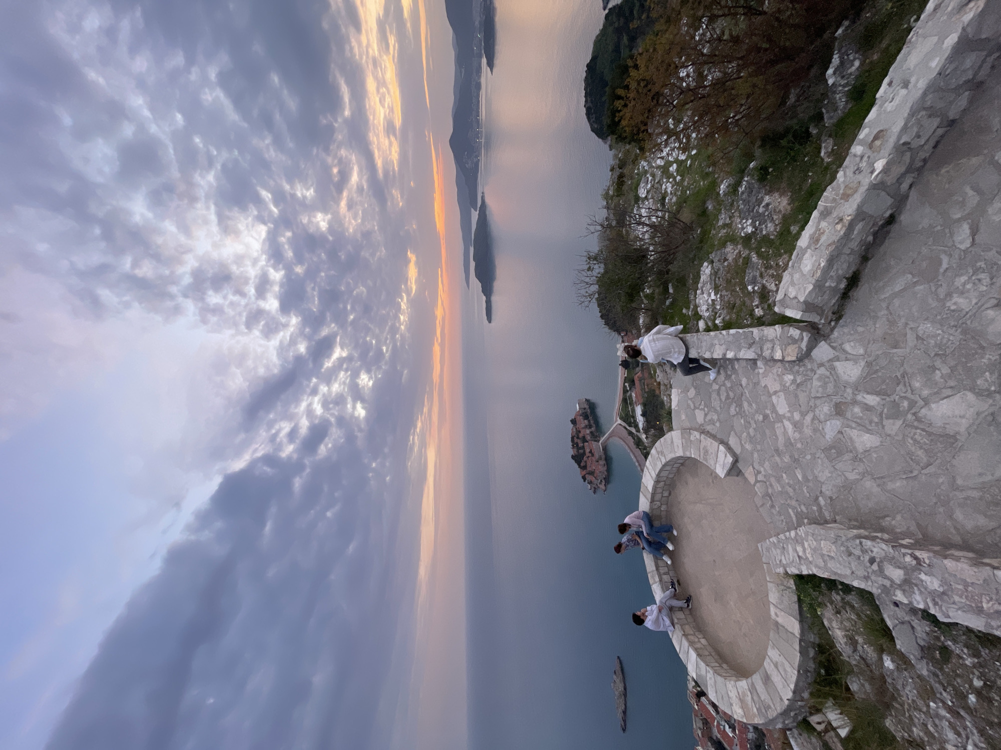 A breathtaking coastal vista, likely captured at sunset or sunrise, shows a dramatic sky with clouds ranging from soft pastels to darker grays, illuminated by a warm, golden glow on the horizon. The image is taken from an elevated stone viewing platform, where four individuals are present.
In the foreground, a curved stone terrace with a low wall serves as a panoramic viewpoint. Two people are seated on this wall, one wearing a white long-sleeved shirt and dark trousers, another in a light-colored top and jeans. Two more individuals stand near the wall, one in a patterned shirt and jeans, and another in a pink top and jeans, observing the scenery. The viewpoint is built into a rugged hillside, with sparse vegetation and rocky terrain visible to the right.
The midground features the calm, reflective waters of the sea, tinted with the sky's soft hues. Prominently centered is the iconic island of Sveti Stefan, with its red-roofed buildings clustered tightly together, connected to the mainland by a narrow causeway. Several smaller islands or rocky outcrops are scattered across the water. In the distance, silhouetted mountains and landmasses stretch along the horizon, fading into the atmospheric haze.
The overall atmosphere is serene and awe-inspiring, with the people seemingly engrossed in the majestic natural beauty of the Montenegrin coastline. There is no visible text in the image.