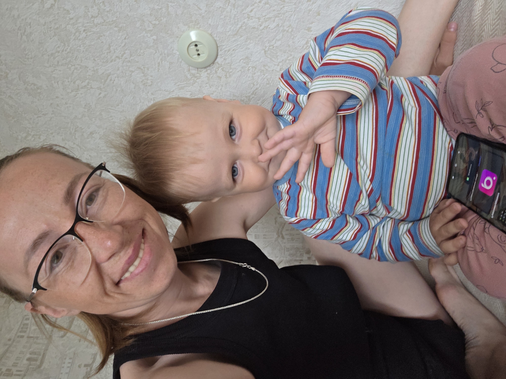 The image is a close-up, upward-angled selfie featuring a woman and a baby indoors. The woman is in the foreground on the left, sporting light brown hair, black-framed glasses, and a black sleeveless top with a thin silver chain necklace. She is smiling broadly towards the camera, her expression cheerful.
Adjacent to her, a baby with light blonde hair and clear blue eyes is visible, also looking towards the camera. The baby is wearing a long-sleeved top with horizontal stripes in blue, red, white, and green. One of the baby's hands is brought up towards its mouth or chin, suggesting curiosity or playfulness. The baby's legs, partially clad in pink patterned fabric, are visible in the lower right section of the frame, indicating it might be sitting on a lap or a soft surface.
The background consists of a light-colored wall with a subtle textured finish. A white, circular electrical socket with two input holes is mounted on this wall in the upper middle portion of the image. In the bottom right corner, a smartphone is visible, its screen illuminated and displaying several application icons. The most discernible icon is a bright pink square with a white, stylized '6' or 'g' symbol. Other icons and text on the screen are too small and blurry to identify clearly. The image conveys a warm and personal moment between the woman and the baby within a domestic setting.