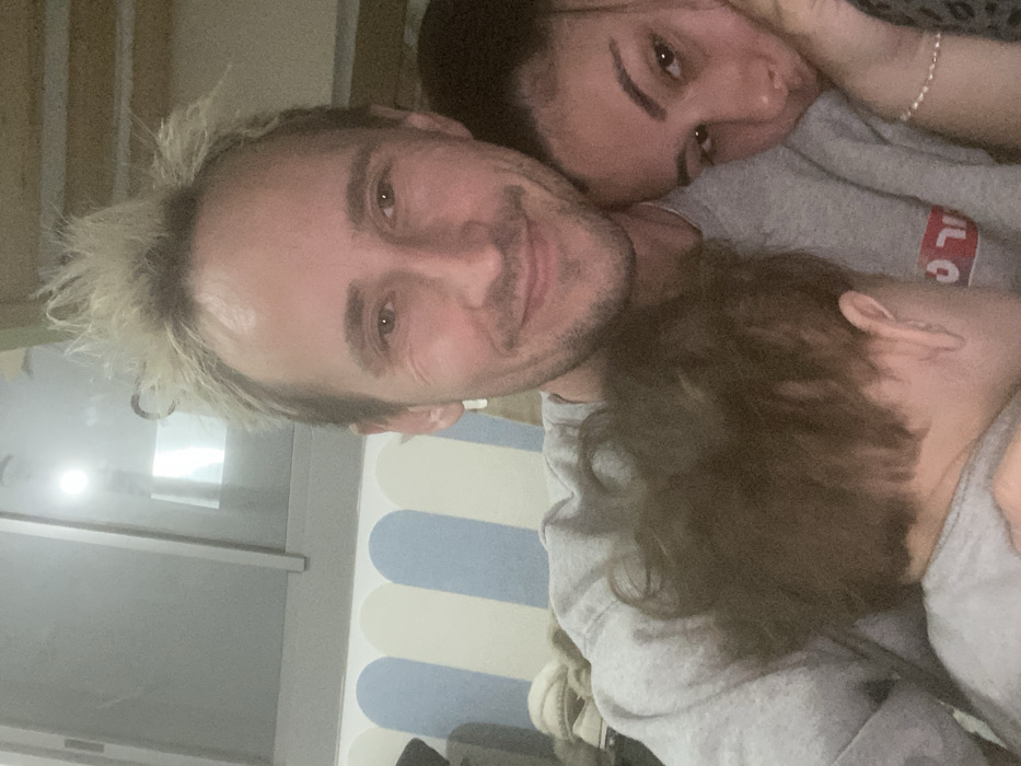 The scene, set indoors in Kiryat Bialik, Israel, features three individuals in a close-up, selfie-style photograph. An adult male occupies the central foreground, smiling directly at the camera. He has bleached-blonde, spiky hair, a light beard, and wears a grey sweatshirt with a partial red and white Hebrew inscription on the chest. A silver hoop earring is visible in his left ear. To his right (viewer's left), an adult female with dark hair, wearing a patterned top and a silver bracelet on her left wrist, rests her head against his shoulder, looking towards the camera with a neutral expression. In the lower-left foreground, a child with dark, curly hair is partially visible, nestled against the man, with the back of their head and right ear facing away from the camera. The background, largely out of focus, shows a light-colored wall adorned with rounded, vertical stripes in light blue and cream, situated beneath a window or glass panel that reflects an interior light source.