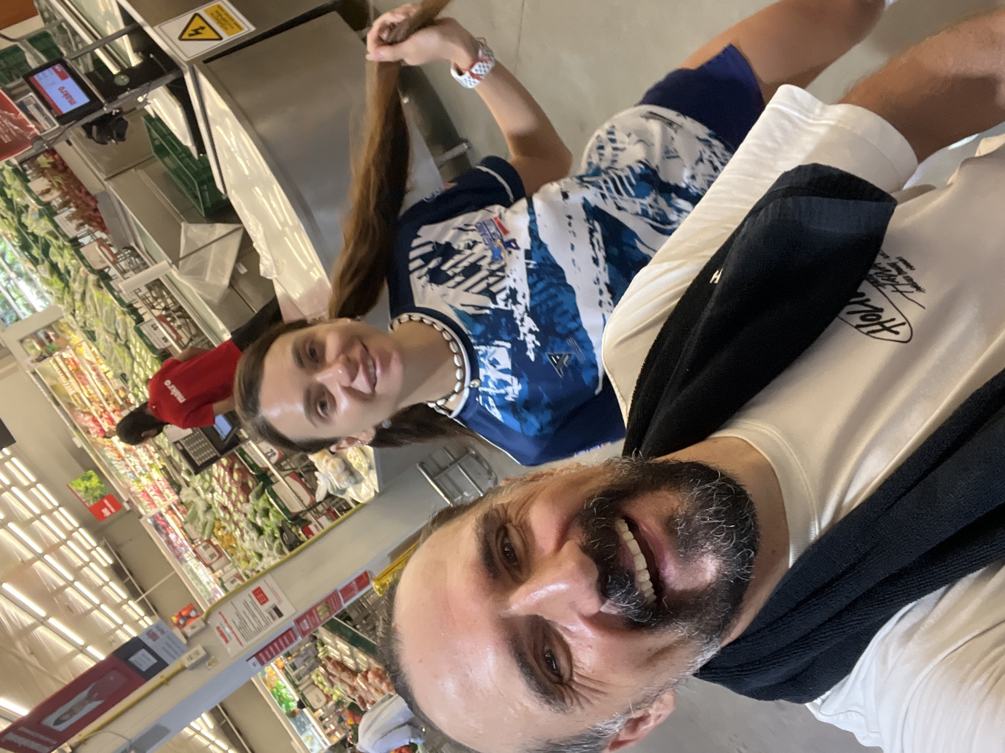 A man and a woman are taking a selfie in a well-lit supermarket in Baan Bo Phut, Thailand. The man, in the foreground, is smiling broadly, with a beard and a black towel draped around his neck. He is wearing a white t-shirt with a graphic print. The woman is behind him, holding her long, dark hair. She is wearing a blue and white patterned t-shirt and a seashell necklace. In the background, shelves stocked with fresh produce are visible, along with checkout counters and other shoppers. The overall atmosphere appears casual and happy.