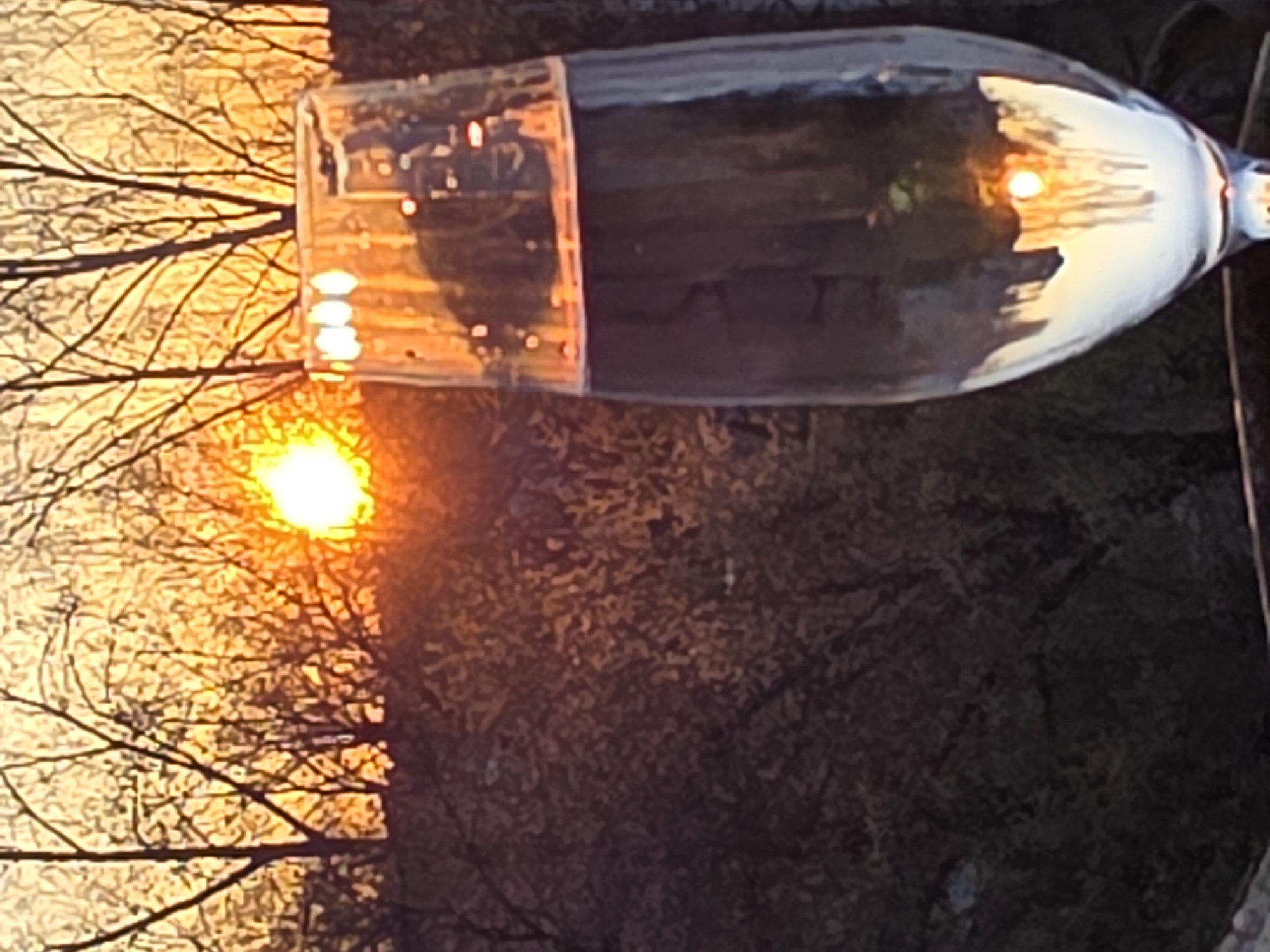 The image shows a sunset over trees, with the reflection of the sunset and trees visible in a partially filled wine glass. The location is likely Chaikovsky, Russia. The environment appears to be outdoors, possibly on a balcony or patio. The primary object is a wine glass reflecting the sunset. The subjects are the sunset and the trees. There is no clear evidence of people.