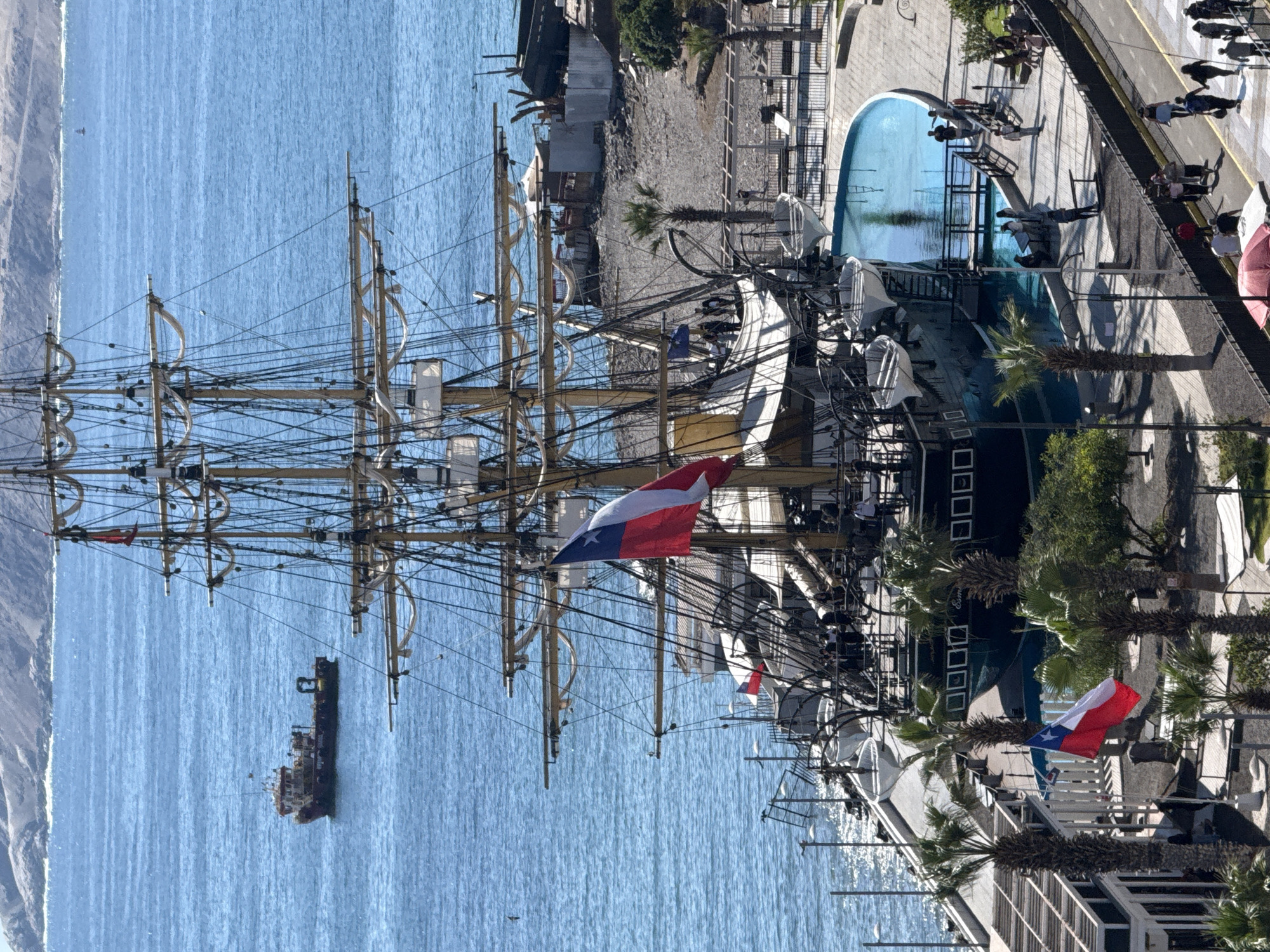 This is an aerial, high-angle, full shot of the Esmeralda training ship docked in the bay of Iquique, Chile. The ship, with its numerous sails and rigging, is the primary subject. Two Chilean flags are visible, one large flag on the ship and a smaller one on a nearby pole. In the foreground, a paved walkway with people strolling is visible, as well as a blue swimming pool adjacent to the ship. The background features a shoreline with buildings and palm trees, and further in the distance, a hazy mountain range and the Pacific Ocean with a small boat. The scene is bathed in bright sunlight, suggesting it is daytime.