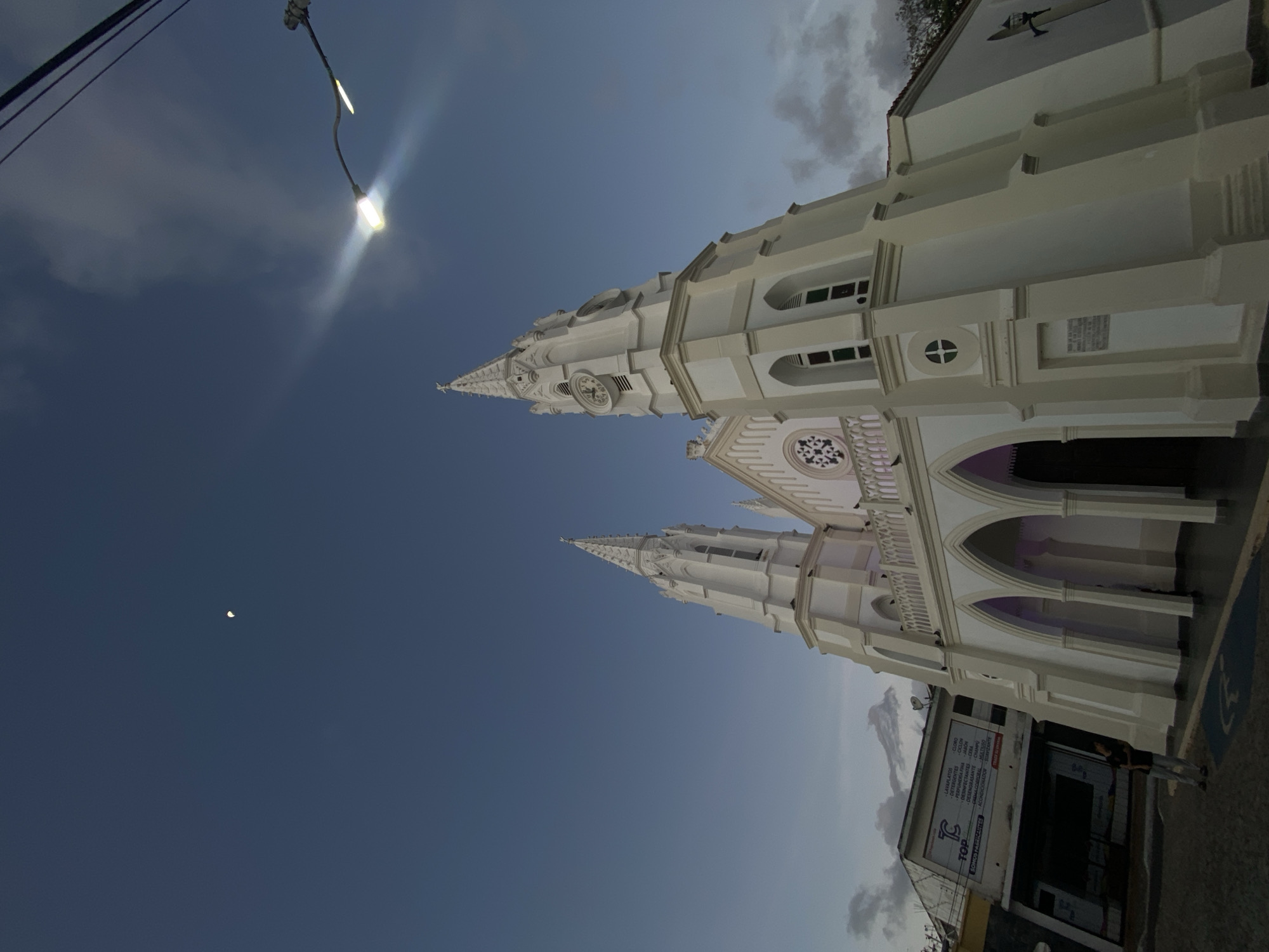 The image depicts the Iglesia de San Juan Evangelista in Juan Griego, Venezuela, at dusk or early evening. The church is a large, white, Neo-Gothic structure with two prominent spires. The left spire is taller and topped with a cross, while the right spire features a clock face. The church facade includes multiple arched entrances illuminated with a purple hue, a balcony with balustrades, and a large circular rose window above the main entrance. Several pigeons are perched on the church's ledges.
To the left of the church, a multi-story commercial building with a sloped roof is visible, displaying a white sign with "TOP" and a list of products in Spanish. A single male individual stands on the paved ground in front of the church, near a blue accessible parking symbol. He wears a dark shirt and light-colored pants. The sky is a deep blue with scattered clouds, and a faint crescent moon is visible in the upper left. A brightly illuminated street light is positioned in the upper right, casting a strong glare.