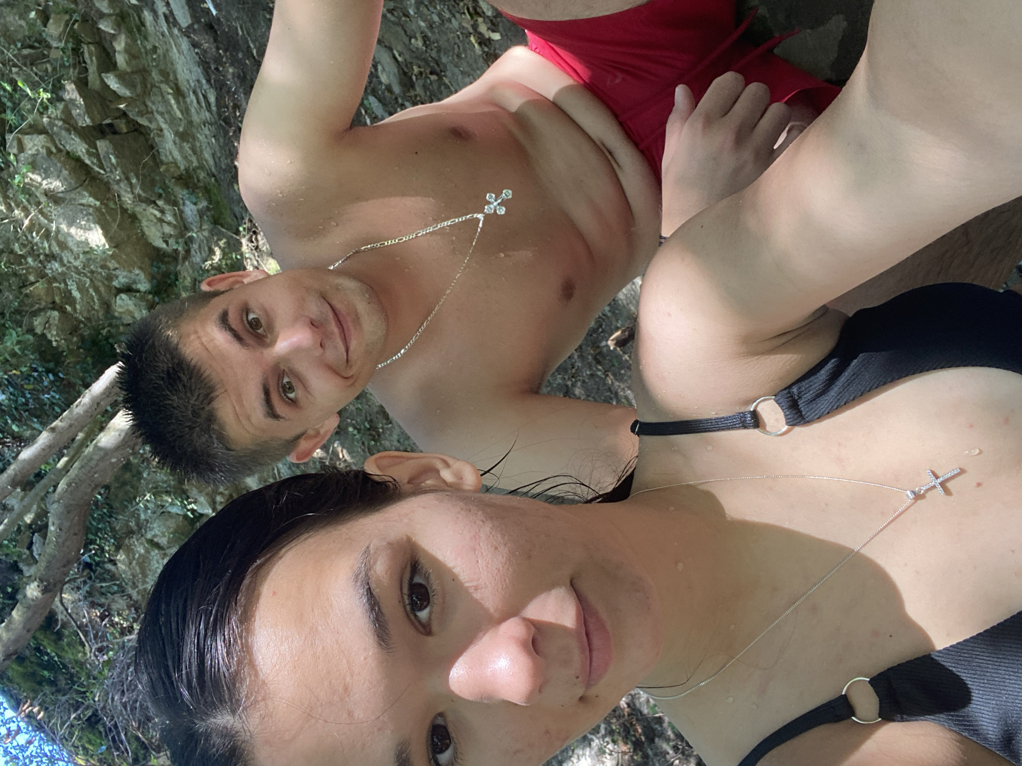 A young couple poses for a selfie outdoors, with the woman in the foreground and the man behind her. The woman, with dark, wet hair and dark eyes, is wearing a black bikini top with silver rings on the straps and a silver necklace with a cross pendant. The man, with short brown hair and brown eyes, is wearing red swim trunks and a silver chain necklace with a cross pendant. They are both shirtless. They appear to be in a natural setting, with rocks and trees visible in the background. The ground is covered in dirt and fallen leaves. There are water droplets on their skin, suggesting they may have been swimming. The lighting is natural and bright, indicating it is daytime. The location is the village of Debnevo, Bulgaria, specifically near a natural water source such as a river or waterfall, which is typical for the area known for its scenic beauty and natural attractions.