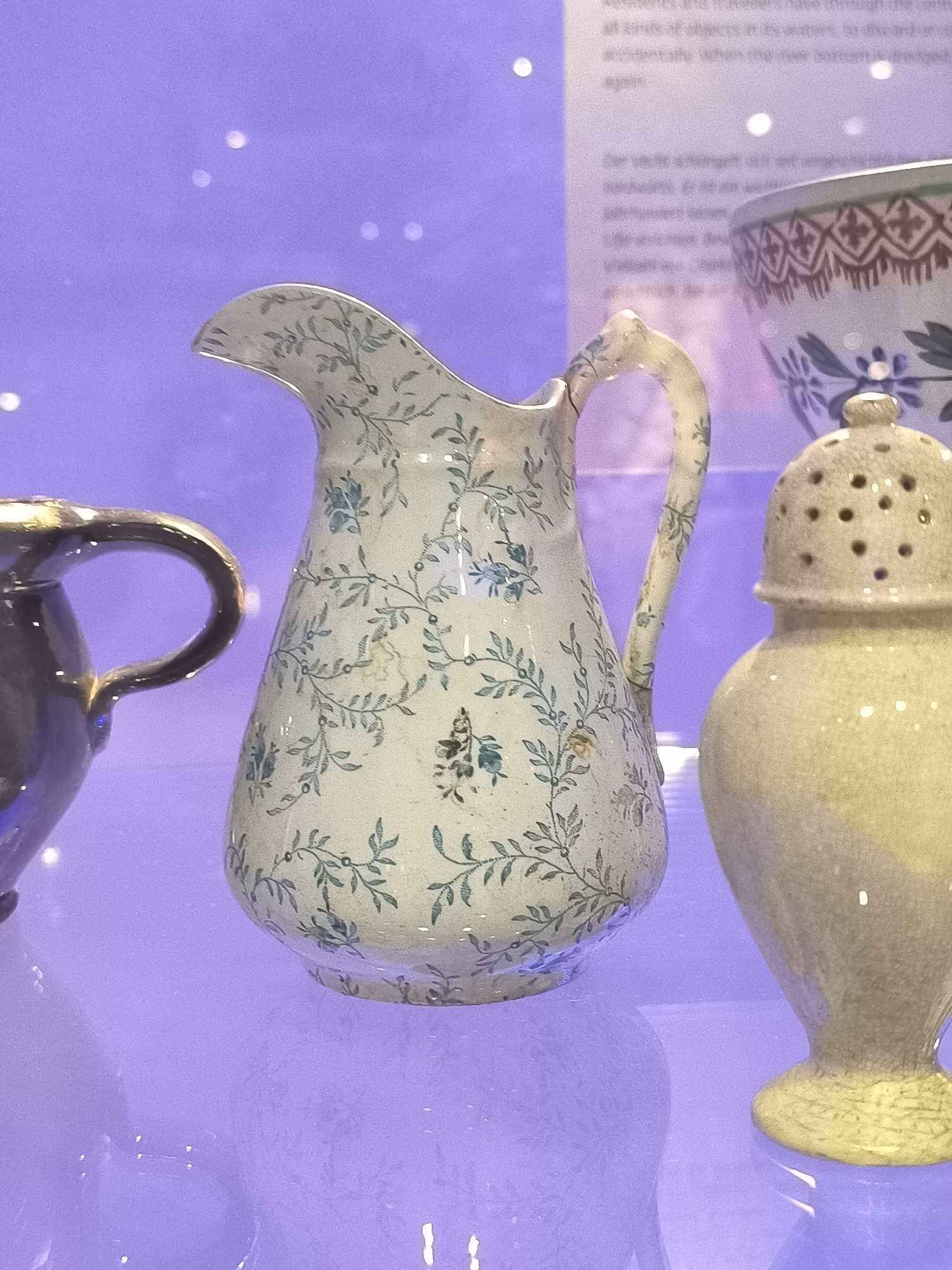 The image presents a still life of antique ceramic pieces displayed within a museum setting. The primary subject is a tall, slender pitcher, primarily white, adorned with a delicate pattern of pale green foliage and small clusters of blue flowers. To the right of the pitcher is a yellow, perforated container with a rounded top, likely a spice or salt shaker. Partially visible on the left is the handle of a dark-colored pitcher. A large bowl with a decorative rim is partially visible in the background to the right of the first pitcher. The objects are arranged on a shiny, reflective surface. Behind the objects, a blurred sign with text in Dutch is visible. Although the image's location within Castricum, Netherlands, is not immediately apparent, the Dutch text on the sign suggests its presence in the area.
