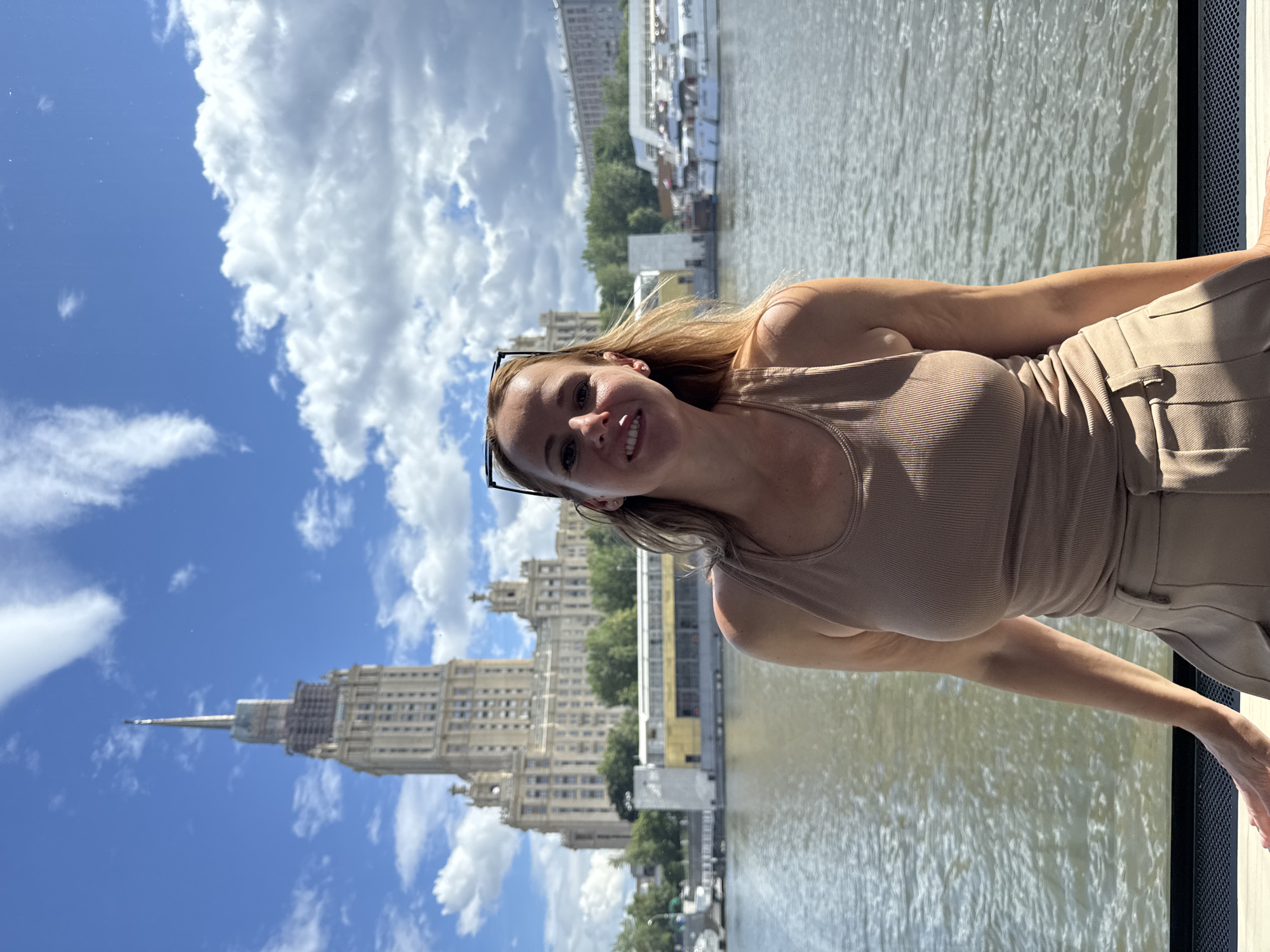 A woman is smiling at the camera, leaning slightly to her left. She has blonde hair and is wearing a tan tank top and tan pants. She is on a boat, with the water of a river visible behind her. In the background, there are buildings and trees, with a tall, ornate building that appears to be a skyscraper with a spire at the top. The sky is blue with fluffy white clouds. The lighting suggests it is daytime, likely a sunny day. The overall mood seems relaxed and happy. No visible text is present in the image.