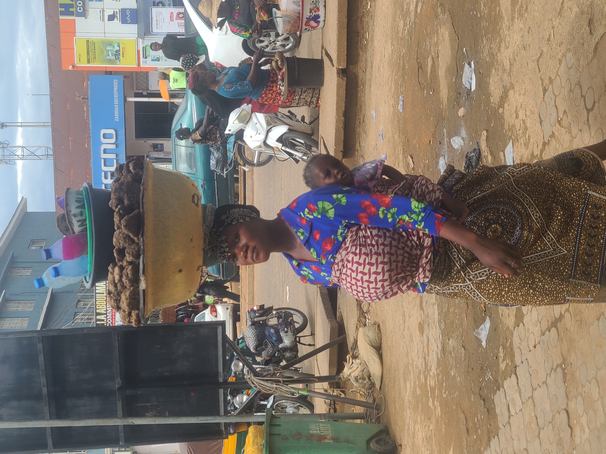 This image captures a daytime street scene in a bustling commercial area within the city of Unknown, Unknown. The lighting suggests an overcast sky or diffused sunlight. In the foreground, slightly right of center, stands a dark-skinned adult female, looking towards the viewer's left with a neutral expression. She wears a blue shirt with red floral patterns, a reddish-brown and beige patterned cloth wrapped around her chest and waist, and a dark brown and beige patterned skirt. Her head is covered by a dark headwrap adorned with small, light-colored decorative elements along the hairline. She carries a stacked load on her head: a large, wide yellow basin filled with dark, lumpy, solid material, and atop that, a smaller dark green basin containing three plastic bottles (one light blue, one dark blue, one pink) and a small purple bowl. A dark-skinned toddler, wearing a light-colored patterned garment, is visible on her back, secured by the chest wrap, looking towards the viewer. The woman's left hand is visible at her side, wearing a ring on her ring finger. To the far left, a green plastic wheeled dumpster, labeled "HAPPY FAMILY" in white, is partially visible. Next to it stands a tall, black metal frame structure, possibly for a billboard or sign. Directly behind this structure, a yellow rickshaw is partially visible. Two motorcycles are parked or standing in the midground left; one is dark-colored with a checkered seat pattern, on a center stand, and another is partially visible behind the black frame. In the midground right, another dark-skinned adult female, wearing a blue patterned top and a red and black patterned skirt, stands behind a dark cylindrical object (possibly a barrel or bucket) serving as a display stand. Green produce, resembling mangoes or avocados, is arranged on top of this stand. A second individual, wearing a straw hat, is partially visible to her right. A white motorcycle is parked near this vendor. The background features a busy street with several vehicles and pedestrians. A light green sedan is visible in the midground, and a white sedan is further in the background. Various commercial buildings line the street. Prominent signage includes a blue sign reading "TECNO" and "ZAKMAN ENTERPRISES," a yellow sign for "NAMAZ COMMUNICATIONS," and smaller signs advertising "VIVO," "HONOR X9A," "LAPTOP," and "HUAWEI." A partially visible sign reads "I.A ABDULMA." Above the buildings, communication towers are visible against the sky. The