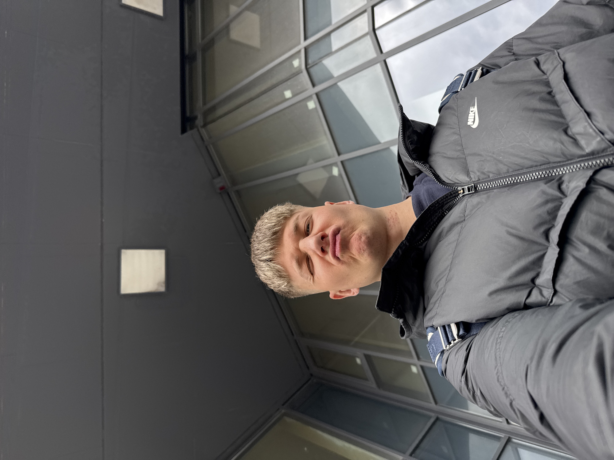 A person with blonde hair is wearing a black Nike puffer jacket while standing in front of a modern building with glass windows and grey walls. The person is making a face with pursed lips. The building appears to be located in an urban environment. There is no indication of this being Plovdiv, Bulgaria.