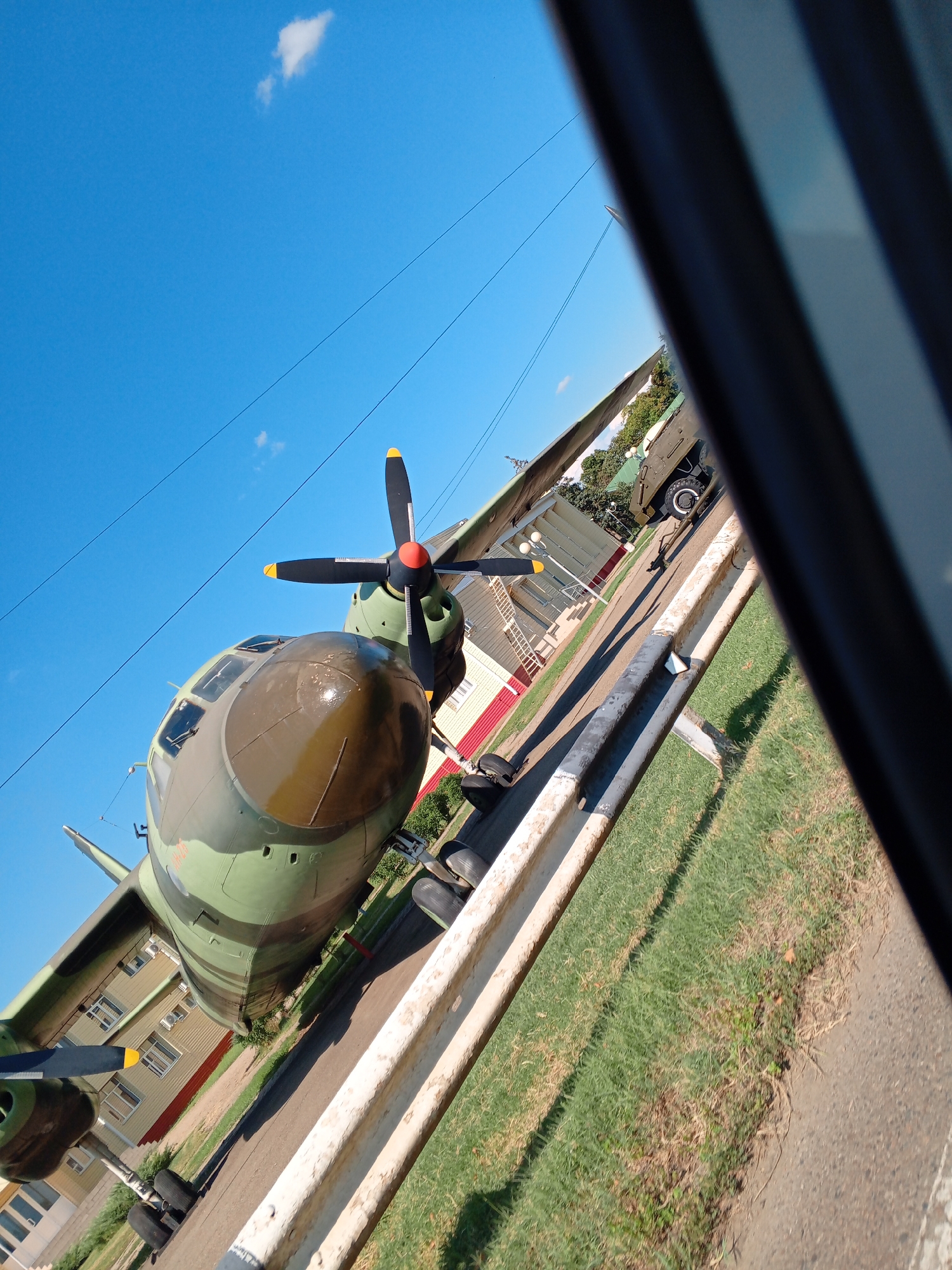 The image shows the front of a camouflage-painted turboprop aircraft parked on an asphalt surface, with a grassy area and a building in the background. The aircraft's propeller is visible, along with its cockpit windows. The sky is clear and blue with a few small clouds. The scene is captured from a low angle, with the aircraft angled towards the viewer. A white metal barrier runs along the right side of the frame, partially obscuring the view of the aircraft. The image appears to be taken outdoors during the daytime.
