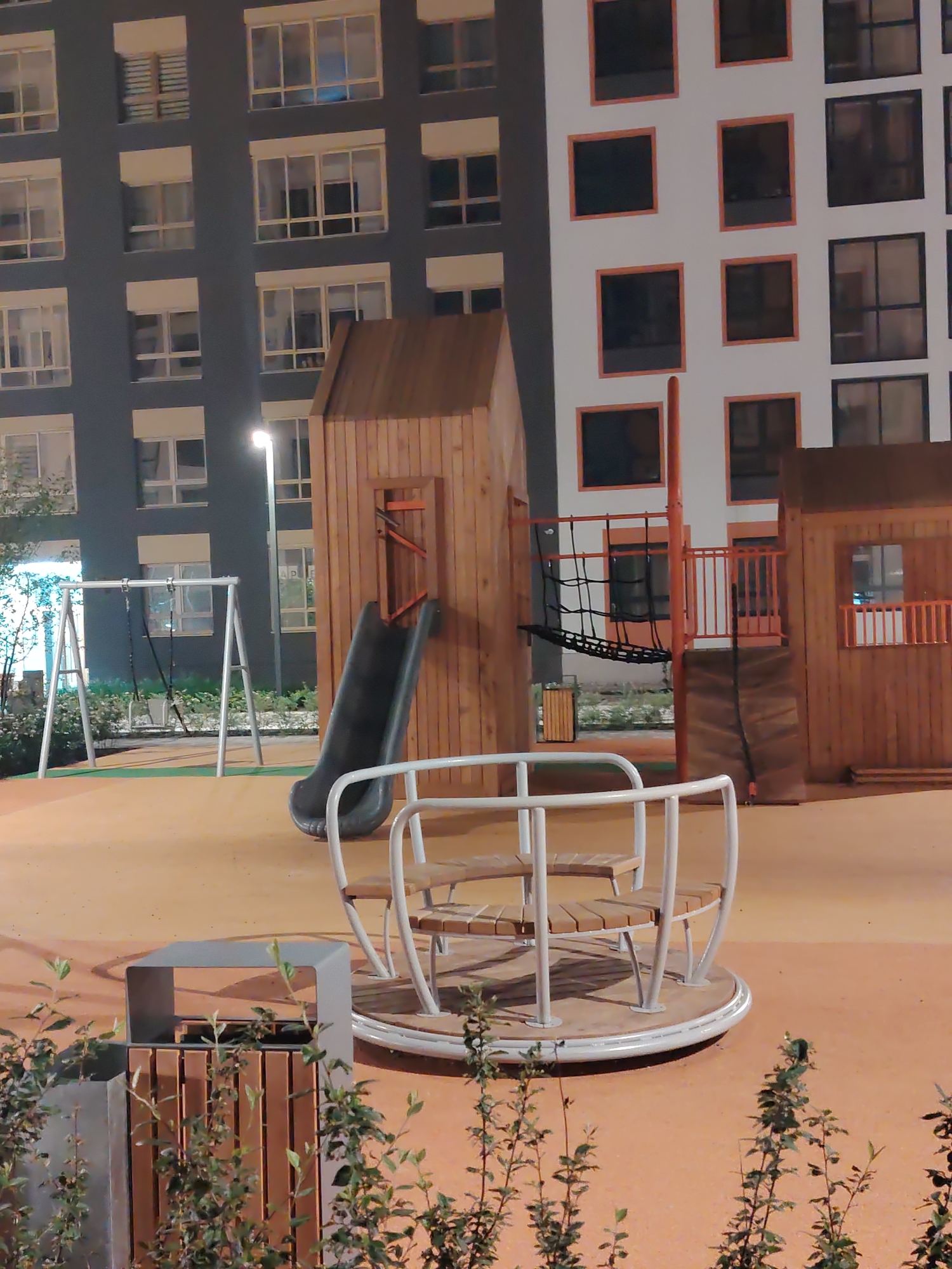 An outdoor playground is illuminated by artificial light at night. The playground features a merry-go-round, swings, a slide, and a climbing net, all constructed from wood and metal. The surrounding area is paved with orange rubber safety surfacing. In the background, two modern apartment buildings rise, one with dark gray cladding and the other with white panels and orange-trimmed windows. The scene is situated in Yekaterinburg, Russia. No people are visible in the image.