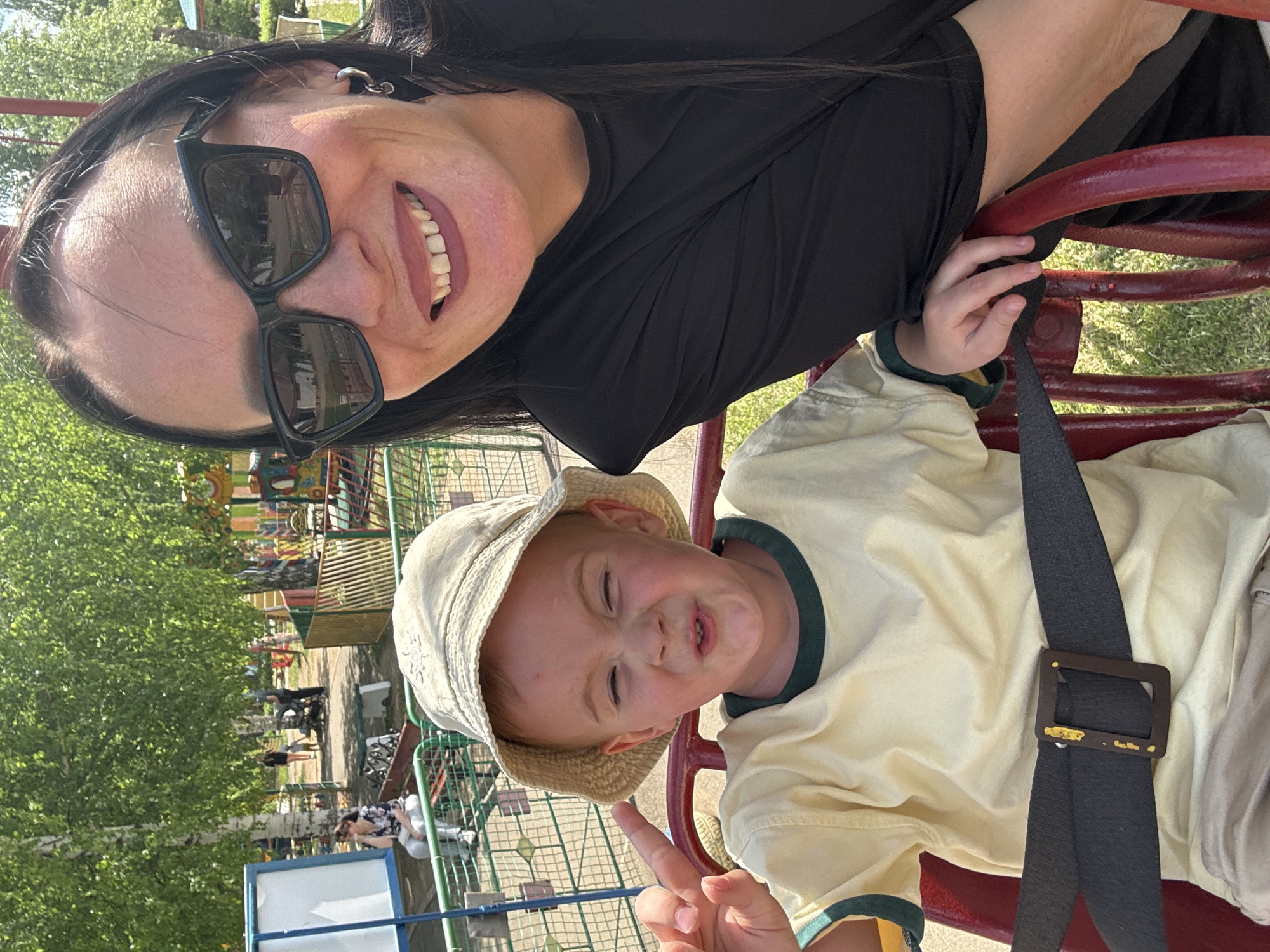 A selfie features a dark-haired woman with sunglasses, smiling, positioned above a child wearing a light-colored hat and shirt. The child is secured in a chair with a black safety belt and makes a peace sign with one hand. They are in a playground setting, identifiable by the playground equipment and trees in the background. The location is Chaikovsky, Russia. Other people can be seen in the background.