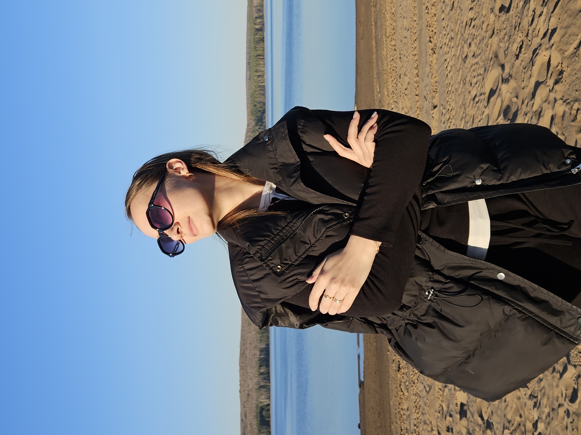 A young woman stands on a sandy beach in Chaikovsky, Russia, possibly near the Kama River based on the water and shoreline visible in the background. She is wearing a black jacket and sunglasses. She has her arms crossed. The sky is blue, and the scene appears to be taken during the daytime.