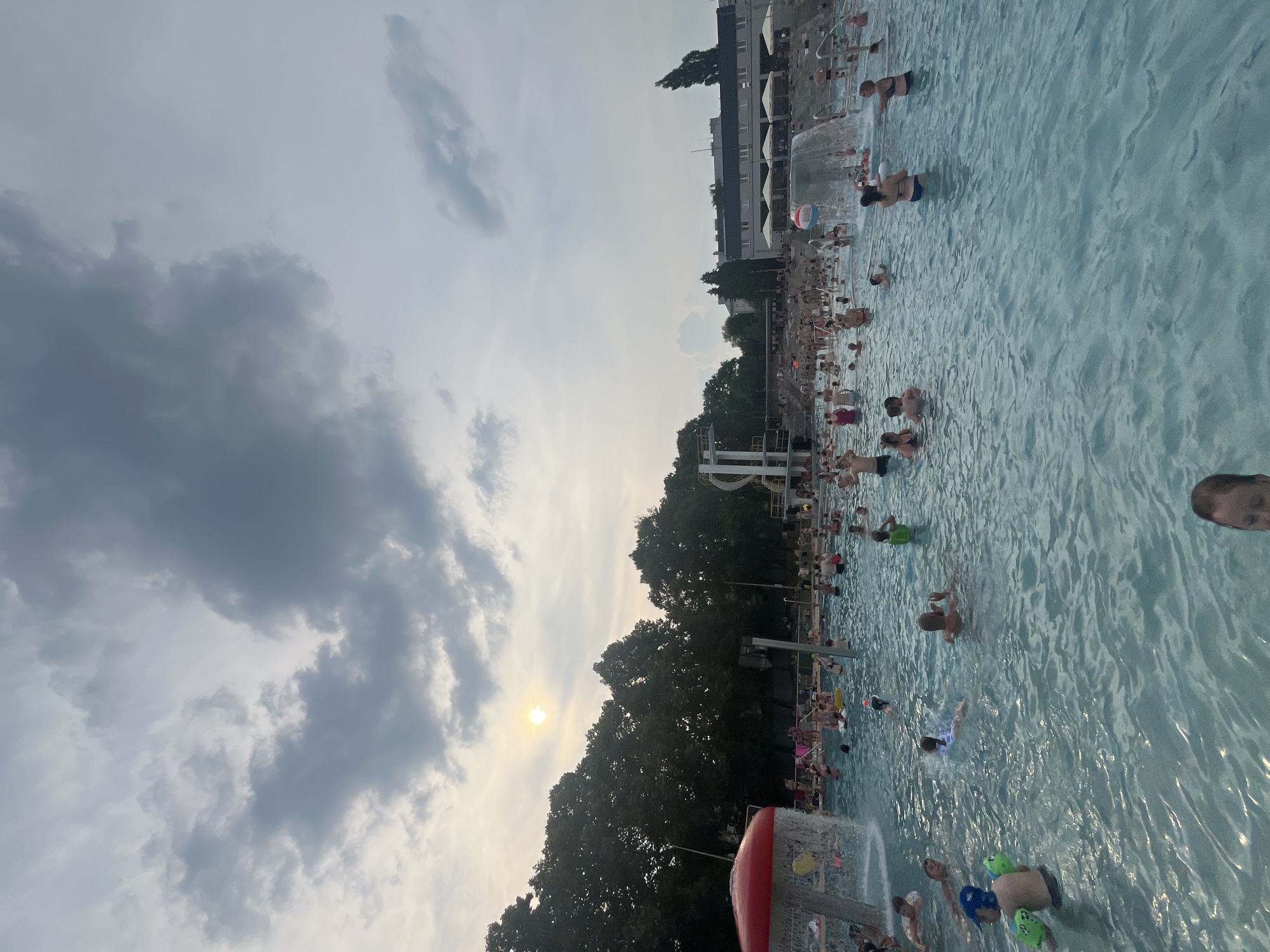 A crowd of people are enjoying a large outdoor swimming pool on a cloudy day. The sun is visible through a break in the clouds, casting a soft glow on the scene. The pool is filled with people of all ages, some swimming, some playing, and others relaxing by the water's edge. There are several features in the pool, including a red mushroom-shaped water feature on the left, and a waterfall on the right. In the background, there are trees and buildings, suggesting the pool is located in a park or recreational area within a city. The overall atmosphere is one of leisure and enjoyment.