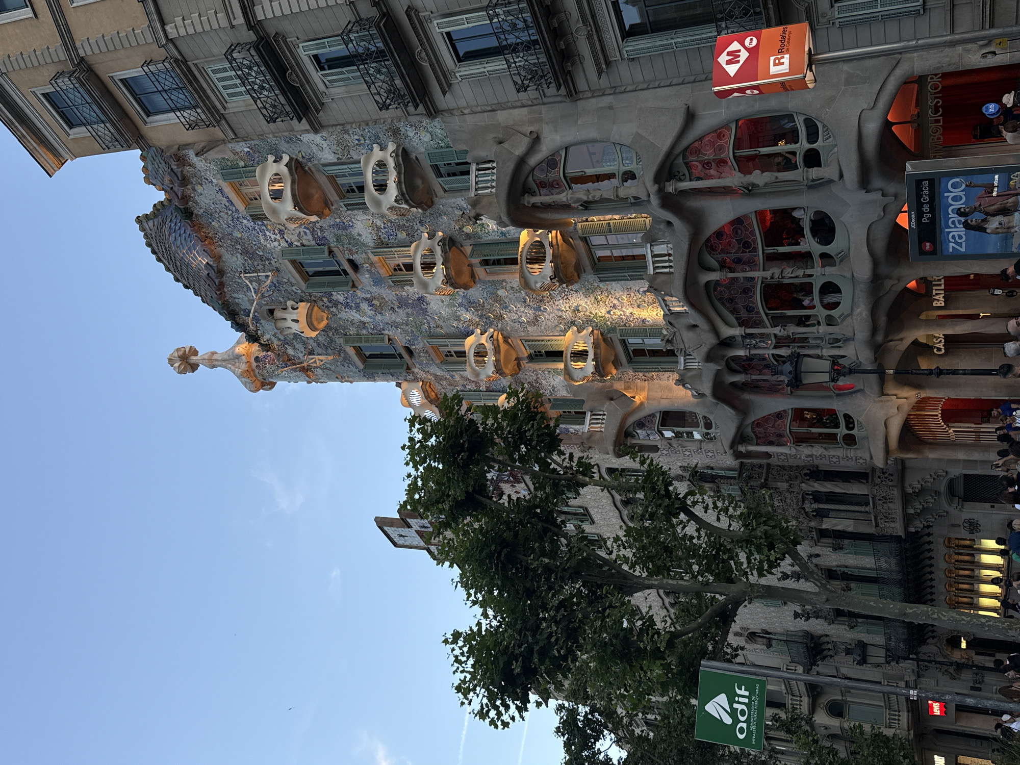 This image, originally rotated 90 degrees counter-clockwise, depicts the iconic Casa Batlló on a lively urban street, likely Passeig de Gràcia in Barcelona, Spain. The unique Modernista building, designed by Antoni Gaudí, occupies the central and upper-left portions of the frame. Its facade is a mosaic of vibrant, scaly tiles, creating an organic, wave-like texture. Bone-like balconies with distinct openings and large, sinuous windows characterize the building's exterior. The undulating roofline, adorned with colorful, patterned tiles and a prominent finial, is visible towards the top.
To the left of Casa Batlló, another building with a more conventional architectural style featuring numerous windows and wrought-iron balconies can be seen. On the right side, the street level of Casa Batlló displays large, irregular window openings resembling caves, with people gathered in front. A row of lush, green-leafed trees partially obscures the lower sections of the buildings and the street.
The scene suggests late afternoon or early evening, as indicated by the clear blue sky with a few faint clouds, the warm light illuminating parts of the building, and the visible illumination from indoor lights and various commercial signs. The weather appears pleasant and clear. People are present on the street and sidewalk, appearing to be tourists or passersby admiring the architecture.
Visible text includes: "CASA BATLLÓ," "Pg de Gràcia" (Passeig de Gràcia), "zalando," "LEVIS," and "SYMBOLIC STOR." Public transportation signs for "M" (Metro) and "R Rodalies de Catalunya" (regional trains) are also present. A green sign reads "adif INFRAESTRUCTURAS FERROVIARIAS," and a digital display shows "21:15."