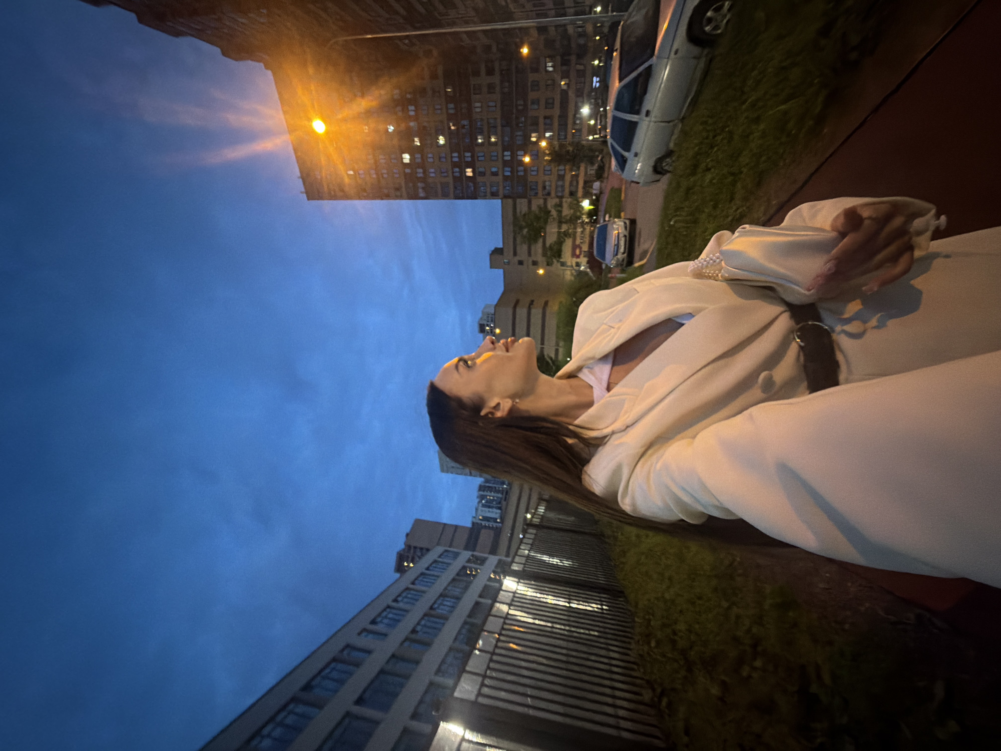 A woman stands in front of a building complex at dusk in Saint Petersburg, Russia. She is looking to the right, her hair flowing over her shoulder. She is wearing a cream-colored blazer with pearl embellishments on the sleeves and is holding a matching clutch. The background features a multi-story building with illuminated windows, a street light casting a bright glow, and parked cars. A fence and grassy area are also visible. The sky is a deep blue, indicating the transition from day to night.
