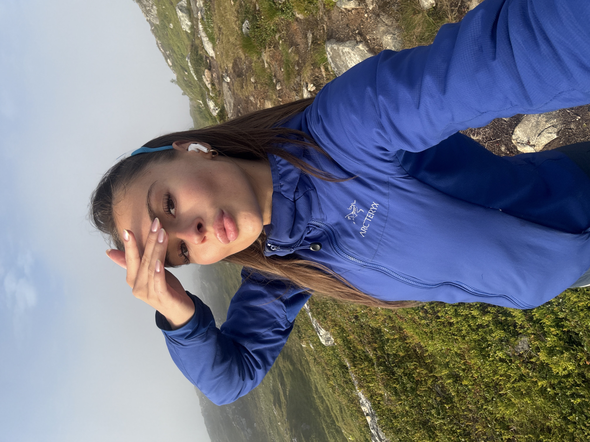A woman is taking a selfie outdoors in a mountainous, misty environment. She is wearing a blue Arc'teryx jacket and white wireless earbuds. Her right hand is raised to her forehead, shielding her eyes from the sun. Her lips are puckered, and she is looking towards the camera. The background shows a grassy hillside with rocks and fog obscuring the distance. The scene appears to be in a natural setting outside of Harstad, Norway, likely during daylight hours given the lighting conditions.