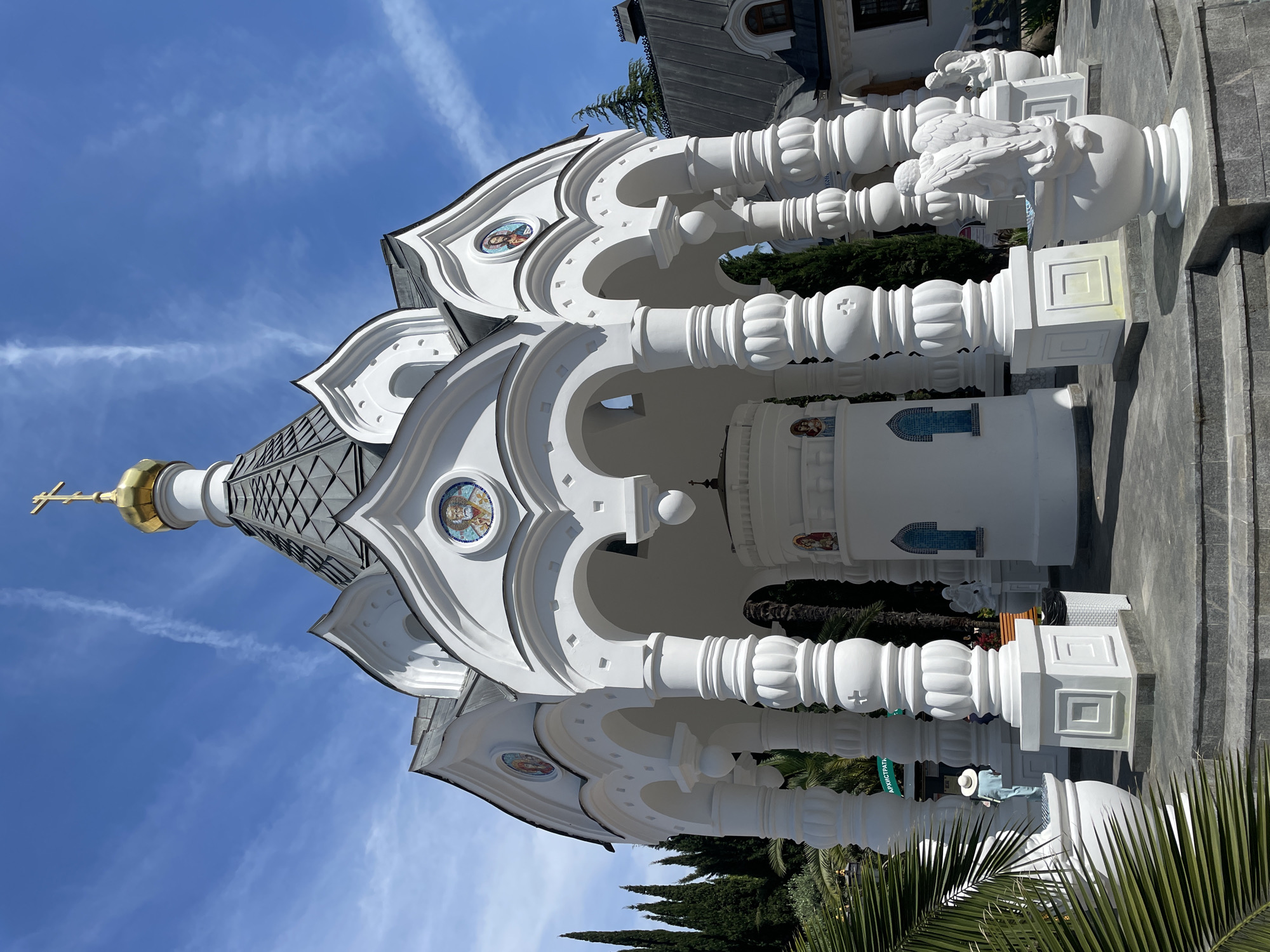 The image presents a low-angle, eye-level exterior view of the white Chapel of Archangel Michael, located in Sochi, Russia, under a clear blue sky with thin, wispy cirrus clouds.
The central subject is an ornate, open-sided chapel, predominantly white, featuring eight prominent, fluted columns supporting a series of arches. Above the arches, a dark gray tiled roof is punctuated by multiple white, decorative, scalloped gables (kokoshniks). Each gable frames a circular mosaic icon depicting a religious figure, with at least four distinct icons visible, including what appears to be Saint Nicholas and Christ Pantocrator. A golden, onion-shaped dome, topped with a golden Orthodox cross, crowns the structure. Inside the chapel's open space, a smaller, cylindrical white structure stands, adorned with smaller mosaic icons and two arched blue mosaic-tile windows or panels.
The chapel rests on a platform accessed by grey steps. The surrounding ground is paved with rectangular grey tiles. To the left, a large green palm frond partially obscures the lower foreground. Behind the chapel and to its sides, tall, slender cypress trees and other green foliage are visible. Part of another building with a dark tiled roof and an attic window is discernible on the far right.
On the left side of the chapel, a white statue of a person wearing a wide-brimmed hat is positioned on a pedestal, partially hidden by the palm frond. A green sign with partially visible Cyrillic text ("АРХИСТРАТ...") is attached to a column base near this statue. On the right side, two white angel statues are placed on white urn-shaped pedestals; one angel faces away from the viewer, displaying prominent wings. A white wicker trash bin is situated near one of the columns on the right. The scene is brightly lit by natural sunlight, casting discernible shadows.