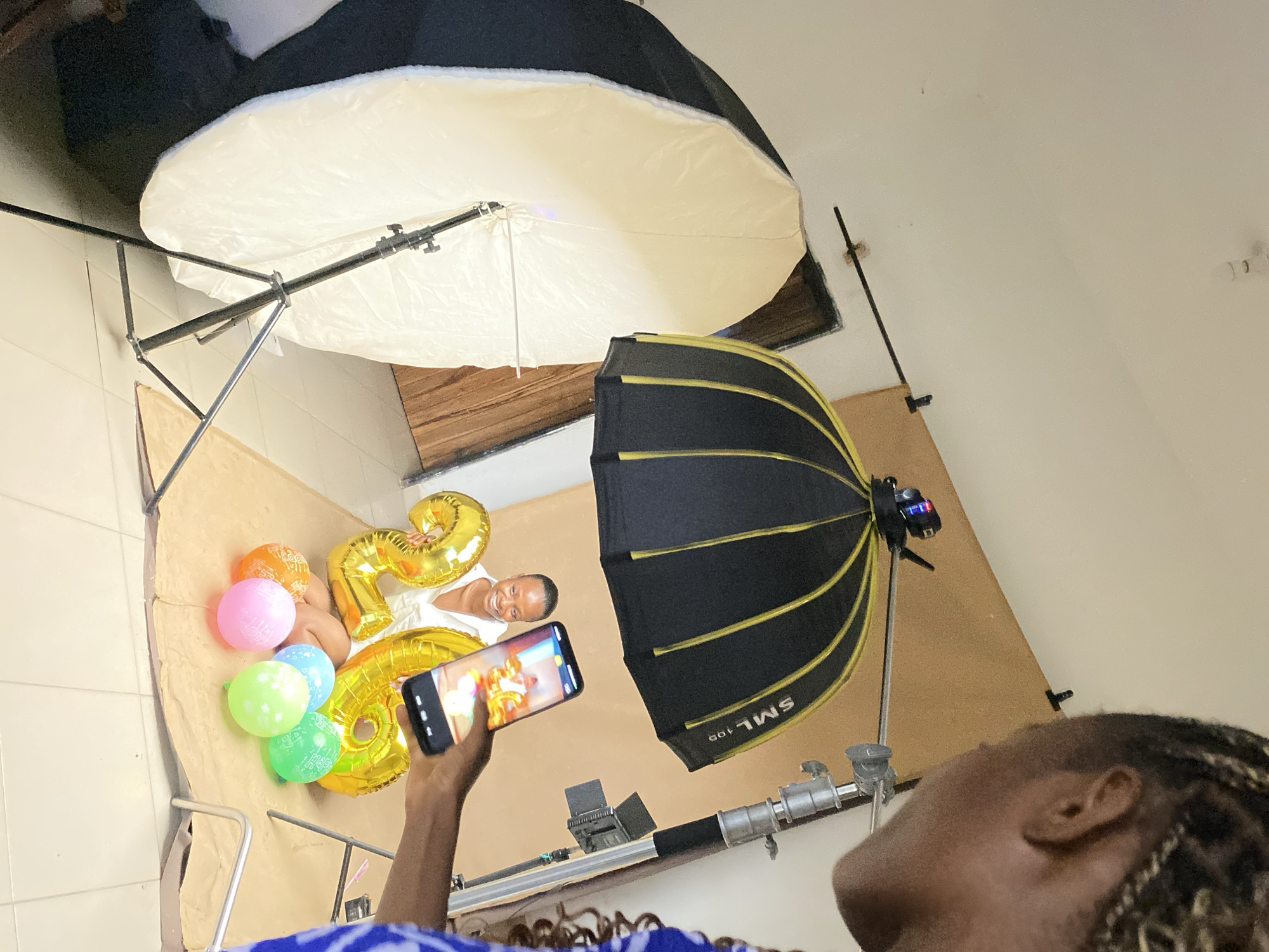 The image shows a behind-the-scenes view of a photoshoot in progress, likely in Jos, Nigeria. A person is lying on a brown surface with gold foil balloons shaped as the number "36" and several colorful round balloons. Someone is holding a smartphone to take a photo of this scene. Two large studio lighting setups are positioned above. One is a large white softbox and the other is a black softbox with gold lining. The environment appears to be indoors, possibly a studio or a room converted into a photo setup. The walls are off-white and there are hints of tiling. The person taking the photo is partially visible, showing their hand and part of their face with braided hair.