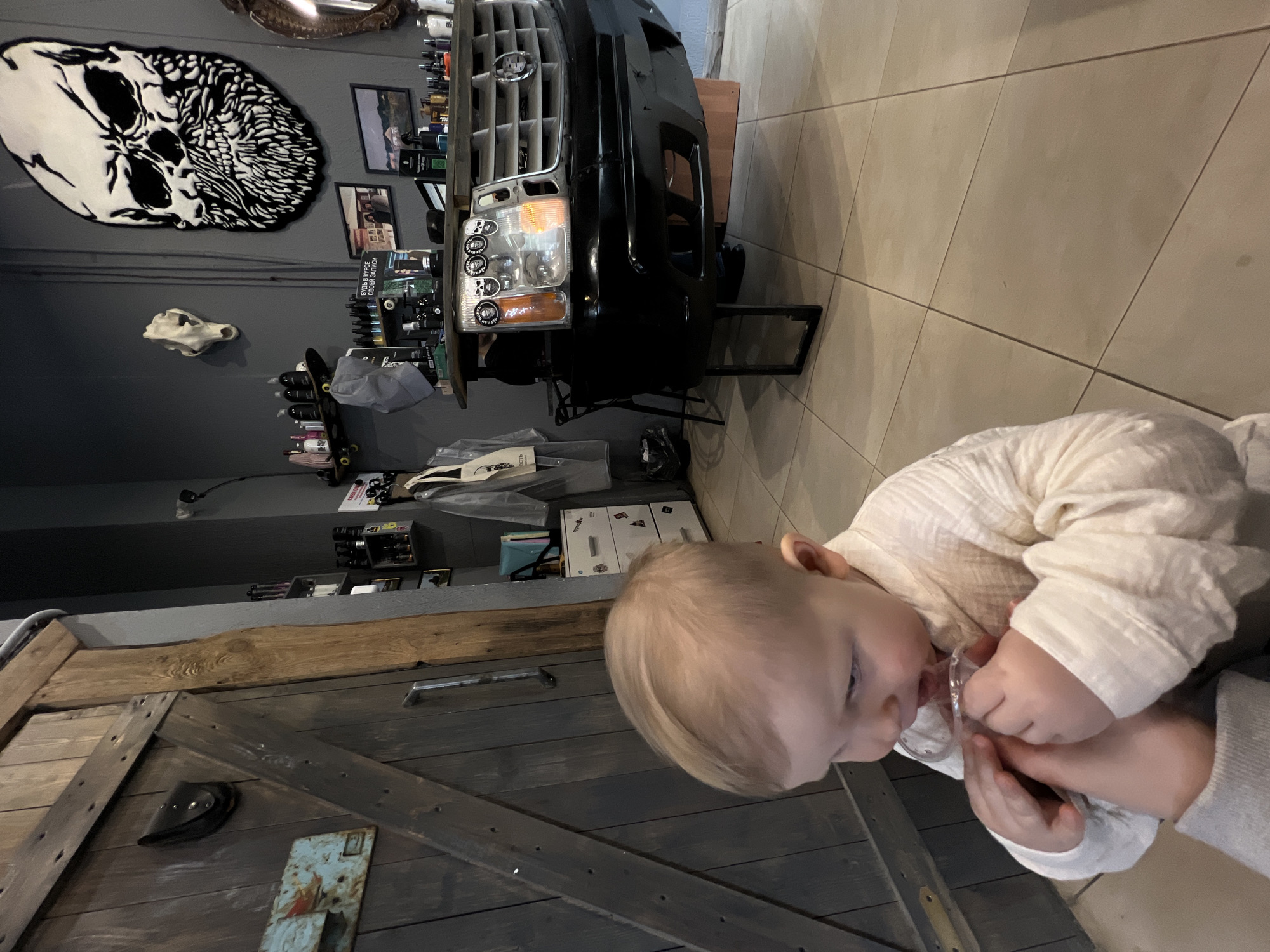 A dimly lit room with gray walls and tiled floors contains a toddler holding a pacifier. The child wears a white shirt. A large wooden door with metal hardware stands nearby. Various items are visible on the walls, including a skull-shaped rug, framed pictures, and a skull replica. A black car bumper is positioned on a table, with a collection of small items and equipment arranged behind it. The location cannot be determined as Khimki, Russia, based on the image alone.