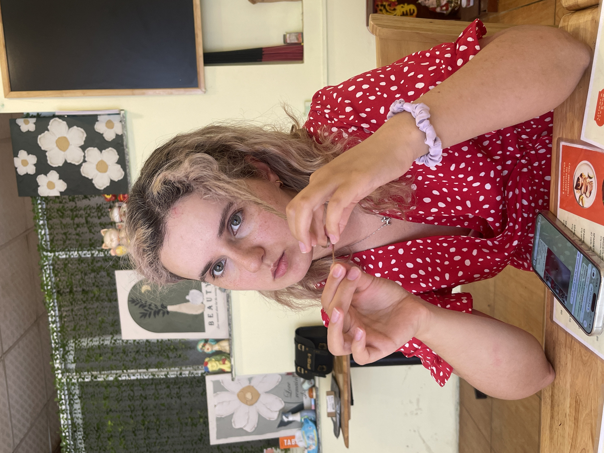 A young woman with blonde curly hair and a red dress with white polka dots sits at a wooden table in what appears to be a restaurant. She has a lavender scrunchie on her wrist. She is holding a strand of hair and looking directly at the camera. A smartphone lies on the table next to her arm. The background features restaurant decor, including artwork with floral motifs and faux greenery. The OCR identifies a menu with prices in Vietnamese currency. The image was taken in Da Lat, Vietnam.
