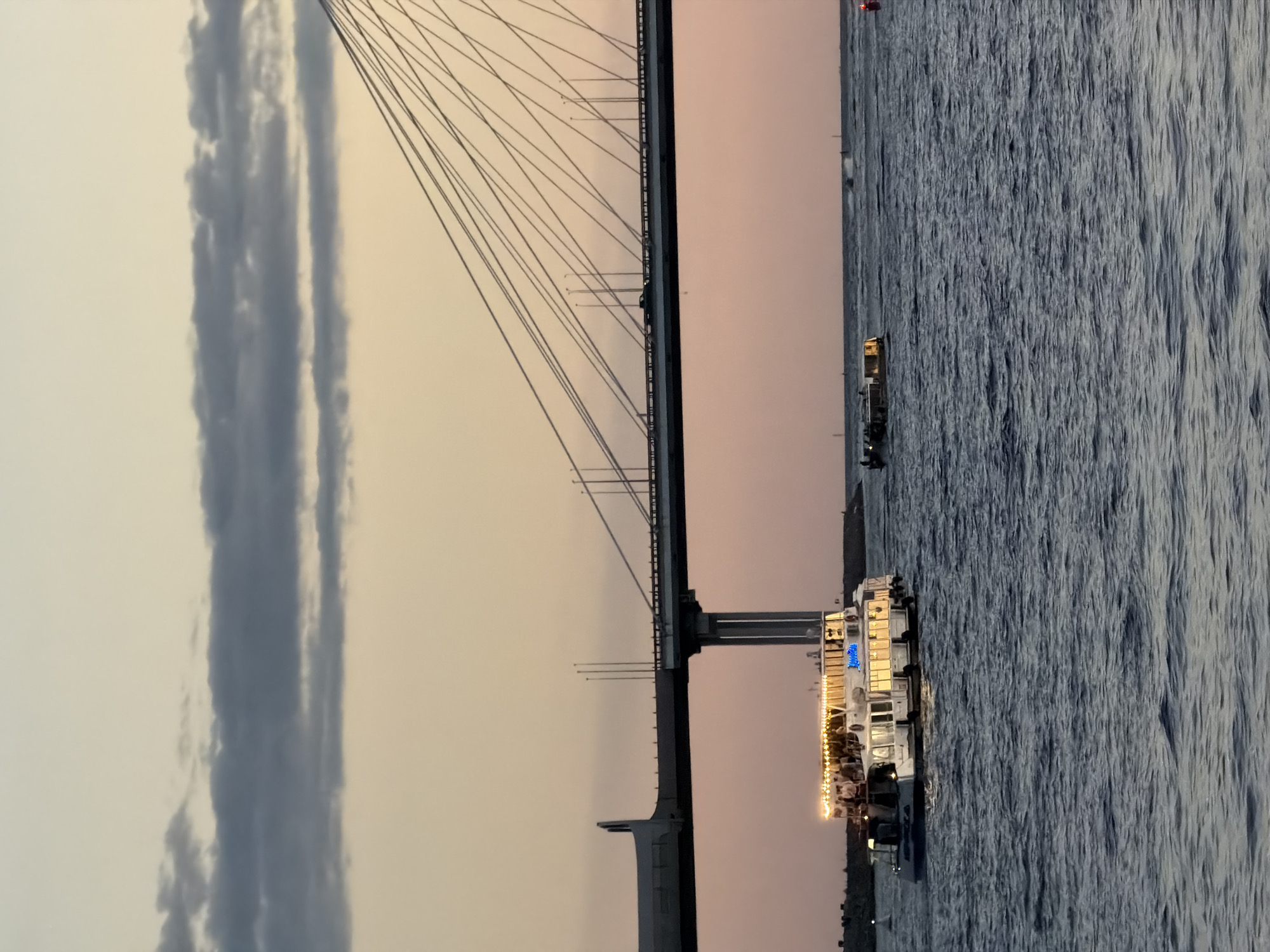 The image depicts a view of the Zolotoy Bridge in Saint Petersburg, Russia, at dusk. A large, multi-deck passenger boat with illuminated windows and string lights is navigating the dark blue water in the foreground, to the left of the frame. The boat has people visible on its upper deck. Further in the background, a second, smaller vessel is also on the water. The Zolotoy Bridge, a prominent cable-stayed bridge, spans across the water, with numerous cables stretching upwards and silhouetted against the pale orange and pink sky. Cars can be seen driving on the bridge, and streetlights illuminate the roadway. The sky is a gradient of soft colors, with some dark clouds visible near the horizon. The overall scene suggests a pleasant evening on the Neva River.
