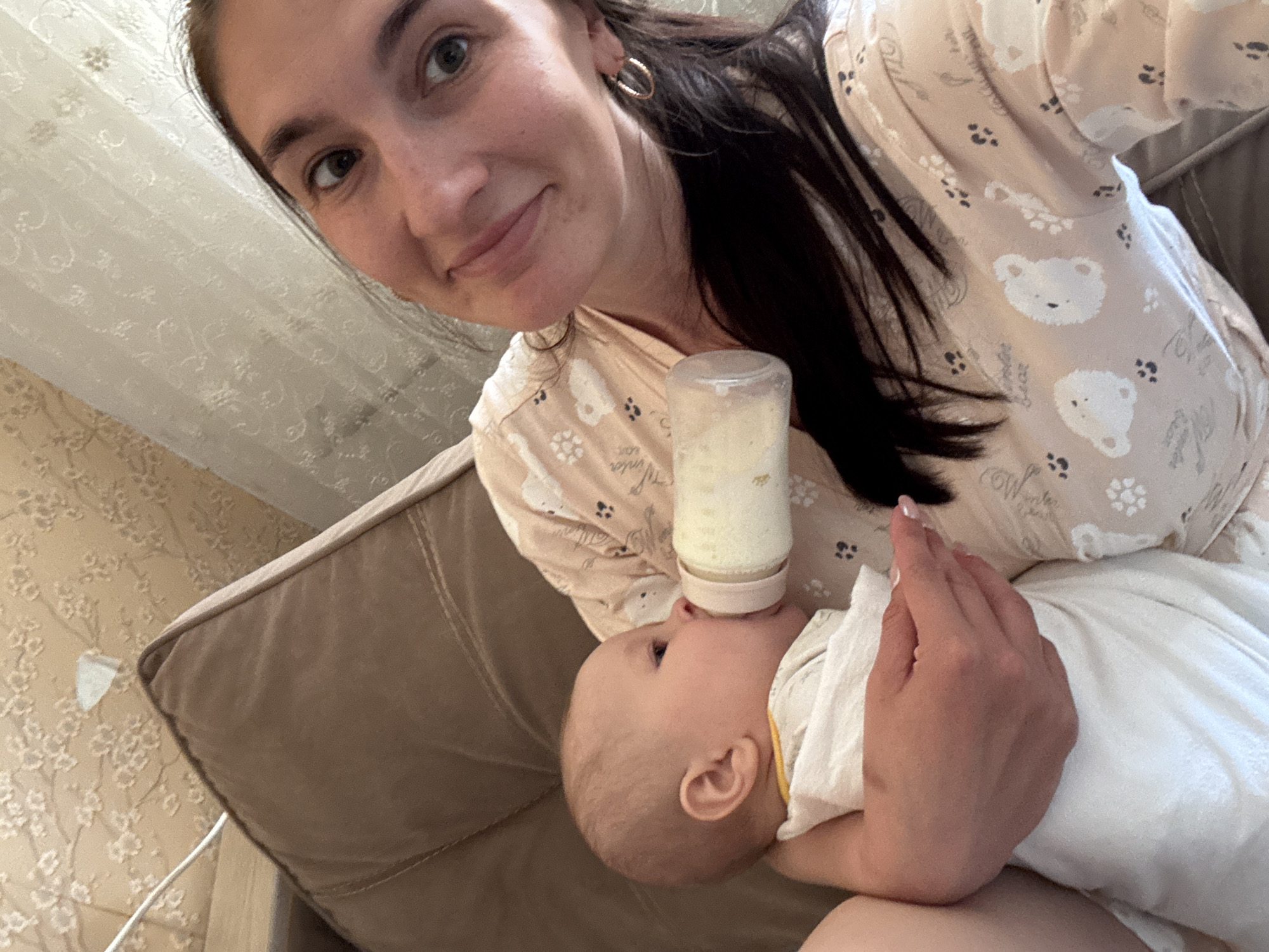 An indoor scene shows a woman bottle-feeding an infant, likely in a living room or bedroom. The image is captured from an elevated perspective, possibly a selfie. The woman, with dark hair pulled back and wearing gold hoop earrings, is positioned on the left side of the frame, looking towards the camera with a soft smile. She wears a light beige top or loungewear patterned with white bear faces, paw prints, and faint text, including "Winter Bear" and "Love".
She holds the baby, who has light hair, in her arms. The infant is lying down, looking up and actively feeding from a clear baby bottle filled with milk or formula. The woman's right hand gently supports the bottle.
The background features a wall covered in patterned wallpaper, possibly with a floral or scroll design, and part of a light-colored curtain or ceiling. A beige couch provides the seating for the mother and baby. The atmosphere is tender and calm, depicting a typical daytime feeding moment between a mother and her child. The location context provided is Sochi, Russia.