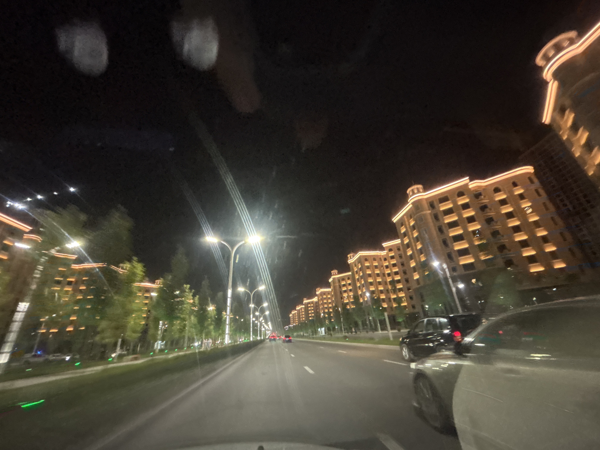 This is a nighttime, low-angle, motion-blurred photograph taken from inside a vehicle looking forward. The scene depicts a wide, well-lit street in the city of Grozny, Russia, lined with multi-story buildings illuminated with warm, orange lights. The buildings appear to have a uniform architectural style, featuring repetitive window patterns and decorative exterior lighting along their roofs and edges.
On the left side of the street, a row of trees with illuminated foliage creates a green and orange glow. Streetlights with multiple lamps arch over the road, casting bright white light. Several vehicles are visible on the road, including a dark-colored car in the foreground to the right, a black car in the middle lane, and red taillights of other vehicles further down the road. The road itself is paved and marked with white lane lines. Small green lights are visible along the grassy median strip. The sky is dark, and there are some indistinct light artifacts, possibly from the camera lens or reflections. The overall impression is a modern, affluent urban environment at night.
