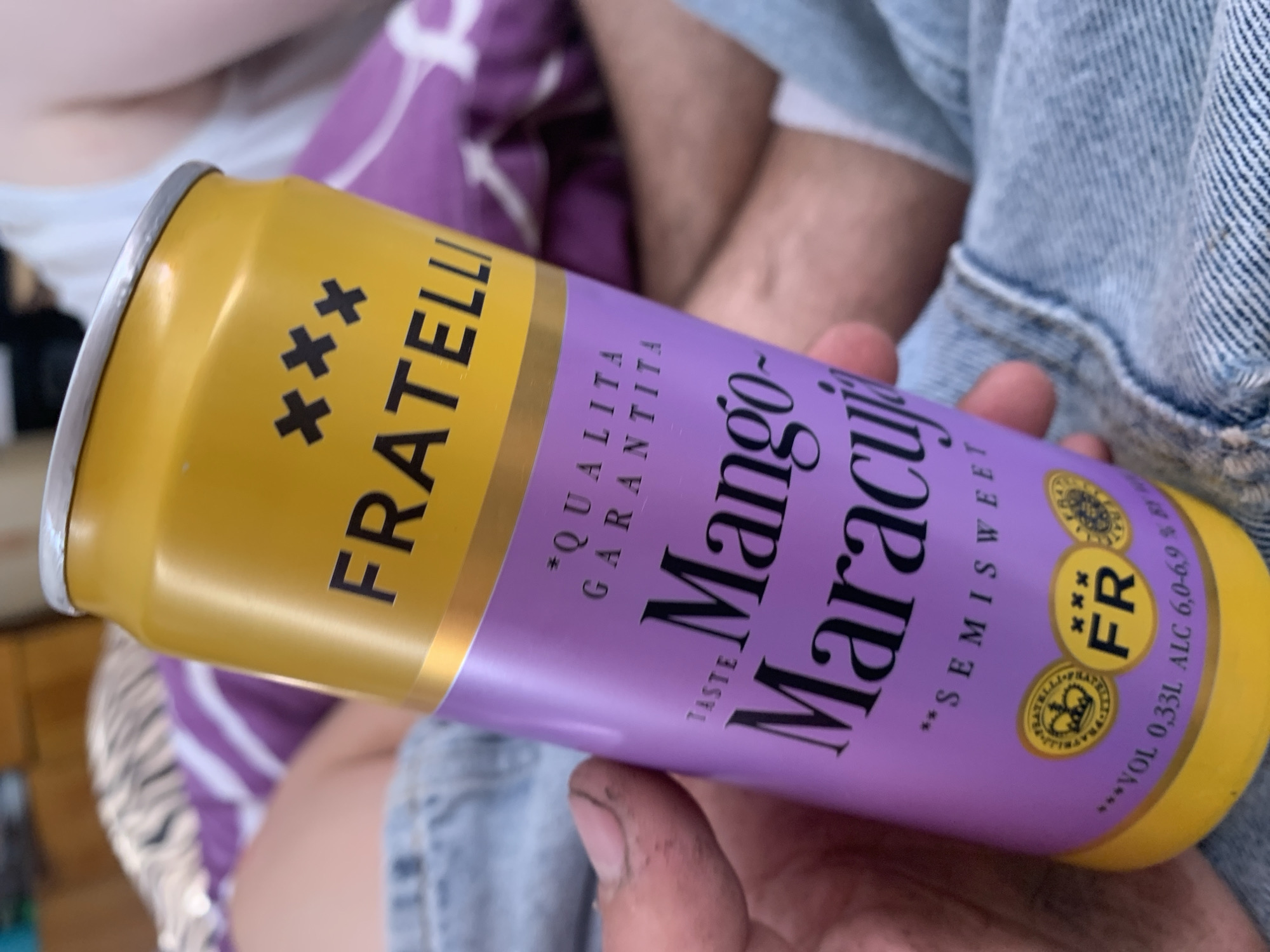 The image shows a close-up of a can of "FRATELLI Mango-Maracuja" drink, held by a person's hand. The can is primarily yellow and purple, with text and logos visible. The background appears to be a blurred indoor setting with what looks like bedding and denim fabric. There is no indication of the city of Zhmerynka, Ukraine, in the image.