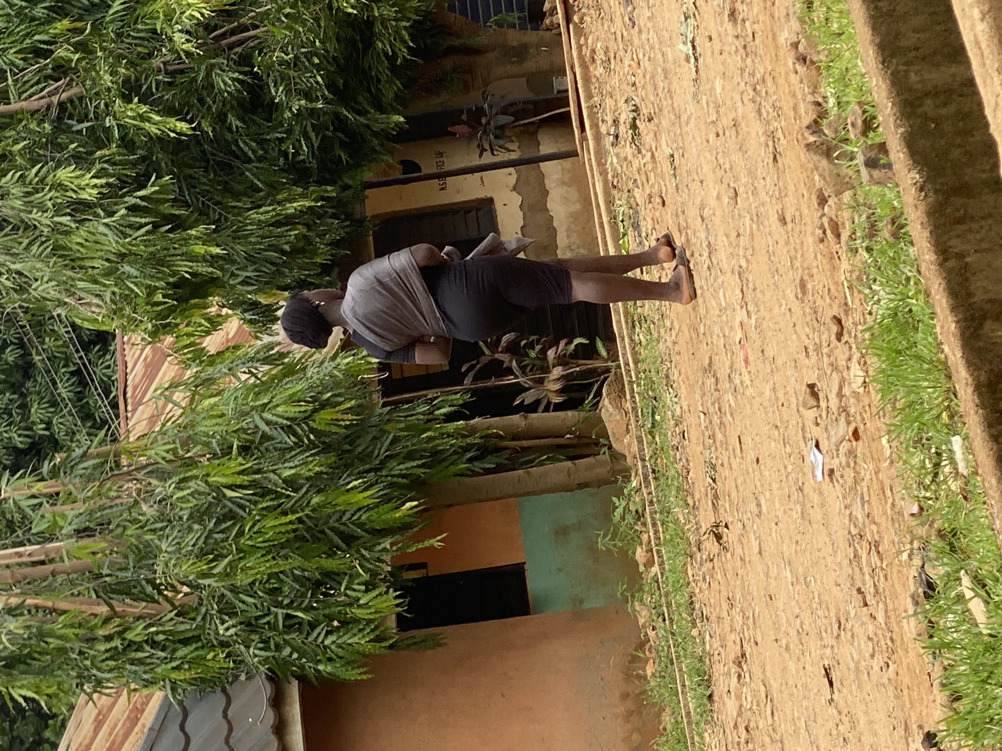 A medium shot depicts a person standing on a dirt path alongside a building in Maigizo, Nigeria. The individual is wearing a gray top and dark shorts, with sandals on their feet, and is holding a document. The building is painted in hues of orange and teal, and is partially obscured by lush green trees. The dirt path is edged with grass. The scene is bathed in daylight.