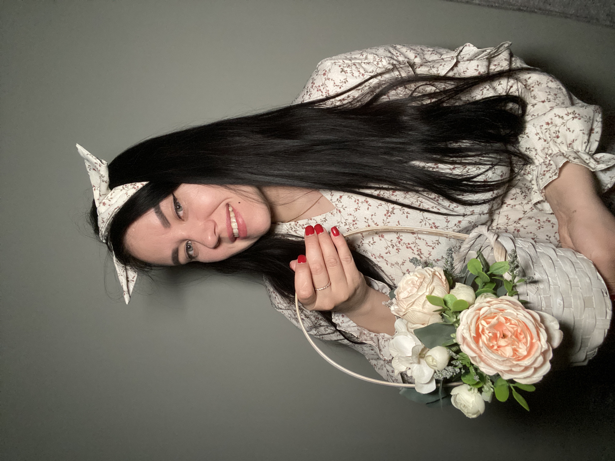 A woman is pictured indoors against a gray background. She is wearing a floral print dress and a matching headscarf with pointed edges. Her dark hair is loosely styled. She holds a white basket filled with cream-colored and peach roses. She is smiling and has red nail polish. There is no information about the location in Chaikovsky, Russia, depicted in the image.