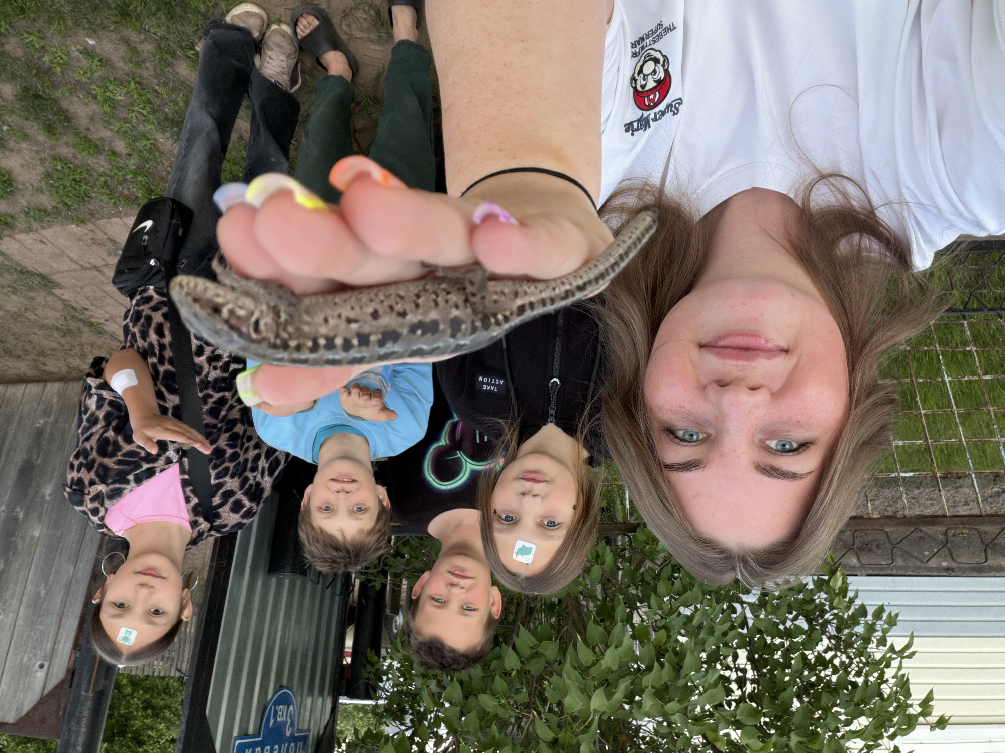 The image captures a group of five people outdoors in what appears to be a residential or backyard setting in Гаревая, Russia. The primary subject is a young woman, visible from the chest up, with light brown hair and blue eyes, wearing a white t-shirt with a "Super Mario" logo. She holds a small, mottled brown lizard between her fingers, showcasing it to the camera. Her fingernails are painted in various bright, pastel colors.
Below her are four children or young teenagers, looking up towards the camera. From left to right: a girl with dark hair wearing a pink shirt under a leopard print jacket and hoop earrings, with a small white sticker on her forehead; a boy with light brown hair in a blue t-shirt; another boy with light brown hair in a black t-shirt (partially obscured); and a girl with dark hair wearing a black zip-up jacket that reads "TAKE ACTION" and also has a small white sticker on her forehead.
The background shows green grass or weeds on the ground, a wooden fence, and a corrugated metal fence. Green foliage from bushes and trees is visible. A blue street sign on the metal fence displays Cyrillic text, including "Гаревая" and "3 кВ. 1". The weather appears to be daytime with soft, indirect lighting. The overall mood is casual and relaxed, with most subjects displaying neutral or slightly smiling expressions, suggesting a moment of shared interest or discovery with the lizard. Visible footwear includes dark sandals and light-colored sneakers. A black cross-body bag with a white Nike swoosh logo is also visible.