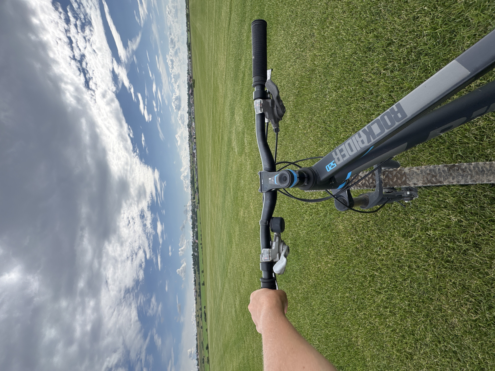 The image captures a first-person perspective from a bicycle handlebar, presumably in Kempelevo, Russia. The scene is dominated by a vast, vibrant green field that stretches towards a distant horizon. A line of buildings, characteristic of a town or village, is visible in the background. The sky above is a dramatic expanse of clouds, with patches of blue sky peeking through. The bicycle itself is a dark color with blue accents, and the name "ROCKRIDER 520" is visible on the frame. A human hand is gripping the left handlebar. The overall impression is one of an outdoor activity, likely a bike ride, in a semi-rural or suburban setting.