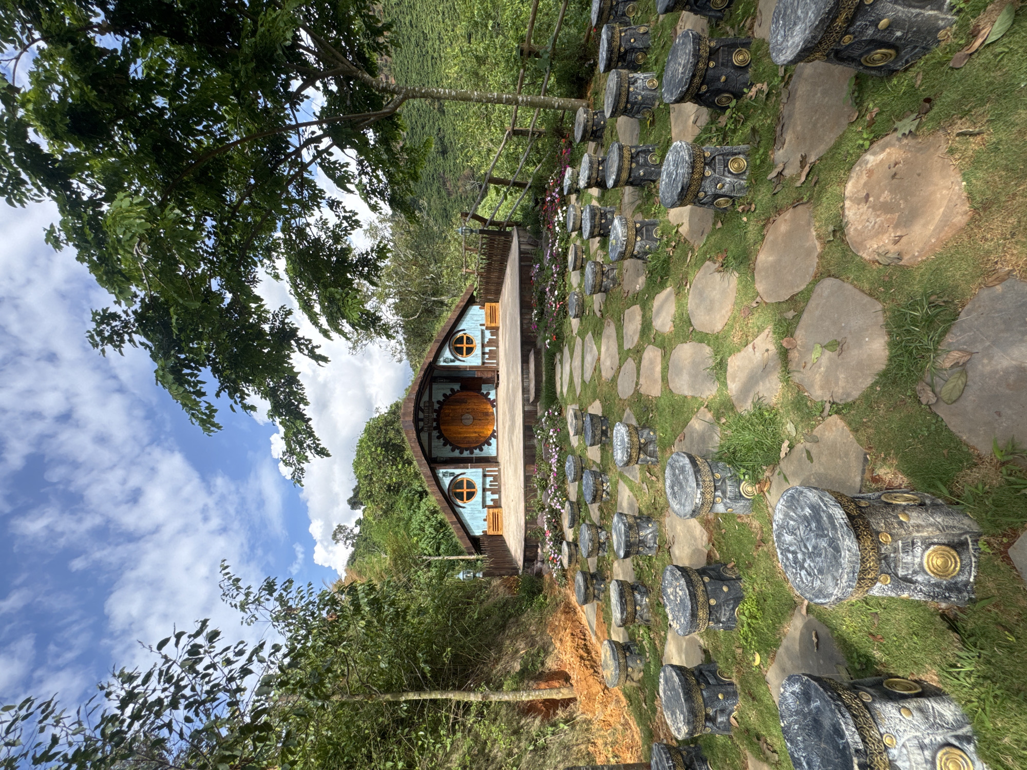 A scene in Thôn 1, Vietnam, depicts a charming, Hobbit-style structure set against a lush, green hillside. The structure, painted blue with a thatched roof, features a large circular wooden door reminiscent of a hobbit hole. In front of the structure, a raised wooden stage is adorned with a colorful array of small flowers. Rows of circular, stone stools, decorated with intricate gold patterns, are arranged on a grassy area interspersed with flat, grey paving stones, suggesting an outdoor seating area or amphitheater. The background is dominated by dense vegetation and trees, with a plantation visible on the slope of the hill. The sky above is a bright blue, scattered with white clouds. No people are visible in the image.