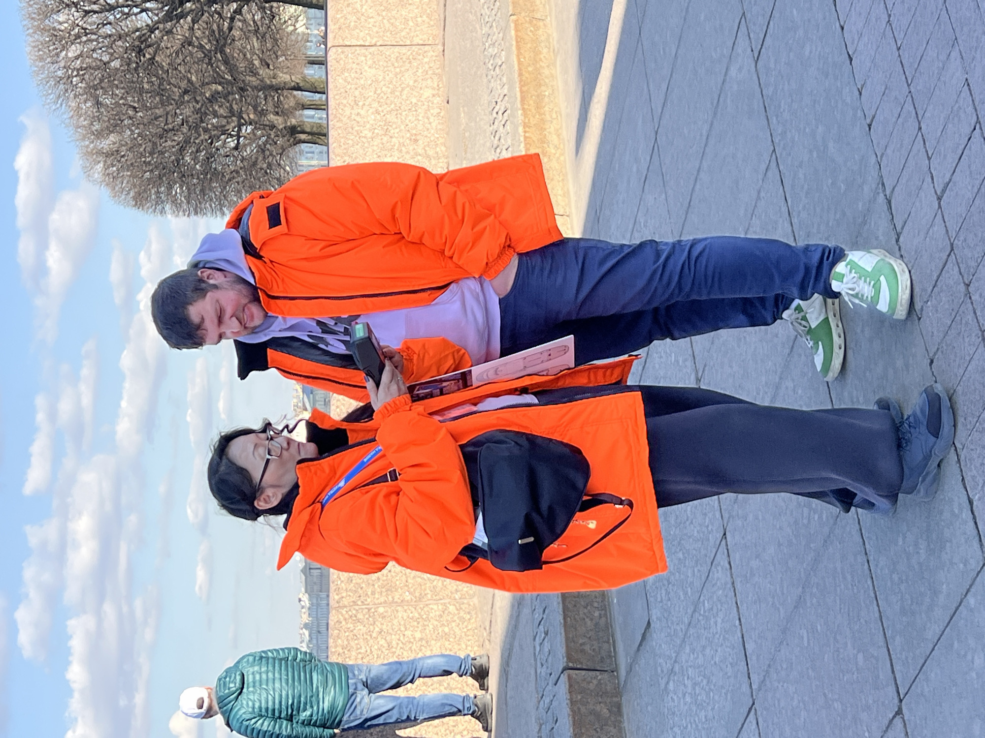 The image captures an outdoor scene on a clear, sunny day in Saint Petersburg, Russia, specifically on an urban embankment. Two primary individuals are in the foreground, standing on a gray paved surface composed of large square tiles.
The female, positioned on the left, appears to be of East Asian descent, wearing glasses and a bright orange puffer jacket over dark wide-leg pants and dark sneakers. A black shoulder bag with a metal accent hangs from her right shoulder, and a blue lanyard is visible around her neck. She holds a dark-colored smartphone or similar electronic device in both hands, looking down at it, and a light-colored, possibly folded, pamphlet or paper in her right hand.
The male, positioned on the right, is a Caucasian individual with a beard, wearing a bright orange puffer jacket over a light purple hooded sweatshirt, dark blue pants, and white and green sneakers. His left hand is in his pants pocket. He holds a dark electronic device, possibly a portable scanner, in his right hand and is looking towards the female and the device she holds, suggesting an interaction or shared focus.
In the far left background, a third Caucasian male is visible from behind, wearing a white baseball cap, a green quilted jacket, blue jeans, and dark boots. He stands near a slightly elevated stone step, observing the distant cityscape.
The environment includes a prominent low wall made of light brown/orange textured stone, separating the paved walkway from what appears to be a lower level or waterway beyond. The distant background features several large, bare deciduous trees, indicative of late autumn or early spring, and a skyline of multi-story classical buildings typical of Saint Petersburg architecture, under a blue sky with scattered white clouds. Sunlight casts distinct shadows on the ground.