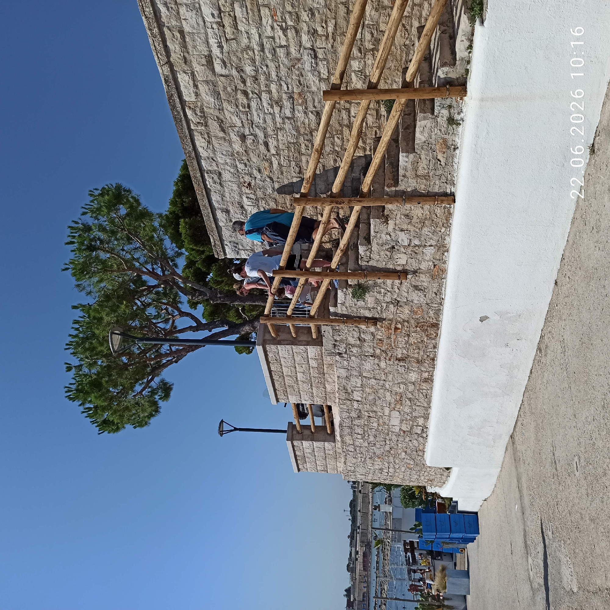 The image depicts a sunny outdoor scene in Trani, Italy, featuring a stone staircase leading upwards along a stone wall. A group of four individuals, two adults and two children, are ascending the stairs. The adults appear to be supervising or walking with the children. A wooden railing lines the staircase for support. At the top of the stairs, near the stone wall, is a tree with green foliage and a lamppost. In the background, a body of water, possibly the sea, is visible with some boats and structures along the shore. The foreground shows a concrete path and some blue crates. The date and time stamp "22.06.2026 10:16" is present in the lower right corner of the image. The architecture suggests a coastal Mediterranean setting.