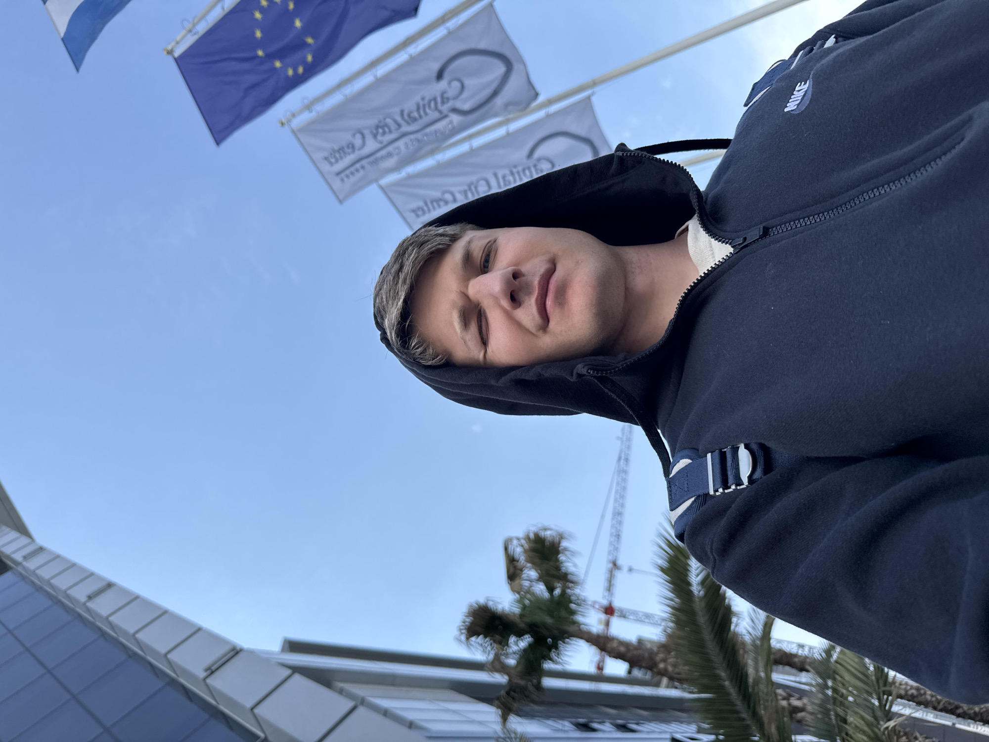 A young man with light skin and short, light-brown hair is the primary subject of this selfie, taken from a low angle looking upwards. He is wearing a dark blue or black hooded zip-up jacket with a white collar visible underneath, and a white "NIKE" logo is visible on the right shoulder area of his jacket. He has a relaxed expression, with a slight smile or smirk and his eyes squinting slightly. A strap or buckle from a backpack is visible over his left shoulder.
The background features a clear blue sky, indicating daytime. In the upper left, three flags are flying. One is blue and white (possibly the flag of Greece), the second is the blue flag of the European Union with yellow stars, and the other two are white flags with a dark logo and text. The text on the white flags, which appears mirrored, reads "Capital City Center" with five stars beneath it, followed by smaller text likely describing a business complex or office area.
Below the flags, to the left, a modern building with geometric glass and metallic panels is partially visible. In the lower right background, several palm trees stand, and behind them, the structure of a large construction crane can be seen, suggesting ongoing development in the area. The overall setting appears to be an urban commercial or business district, consistent with the "Capital City Center" text on the flags.